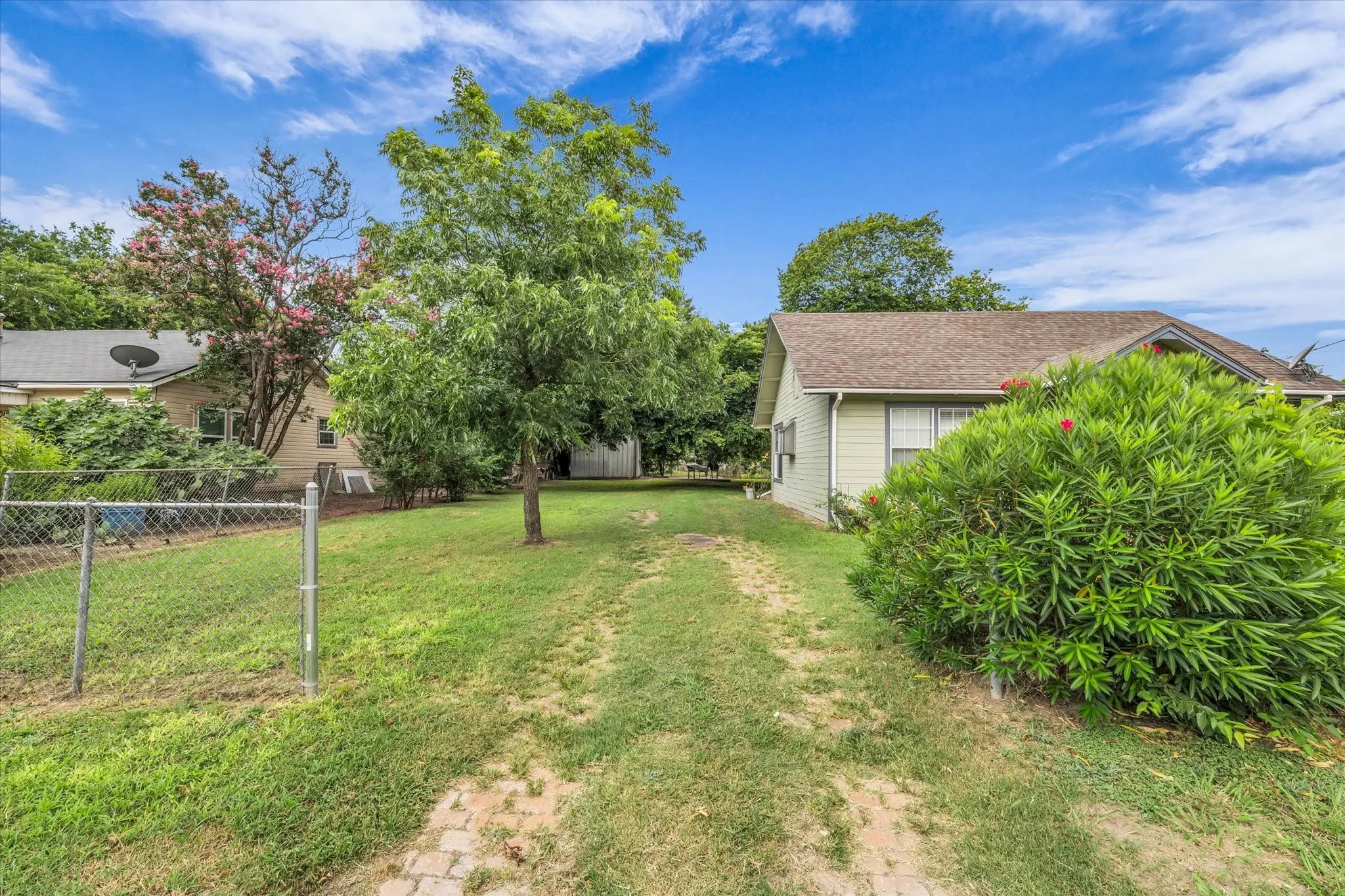 Expansive lot featuring a chain-link fence, mature trees, and a gravel and paver pathway