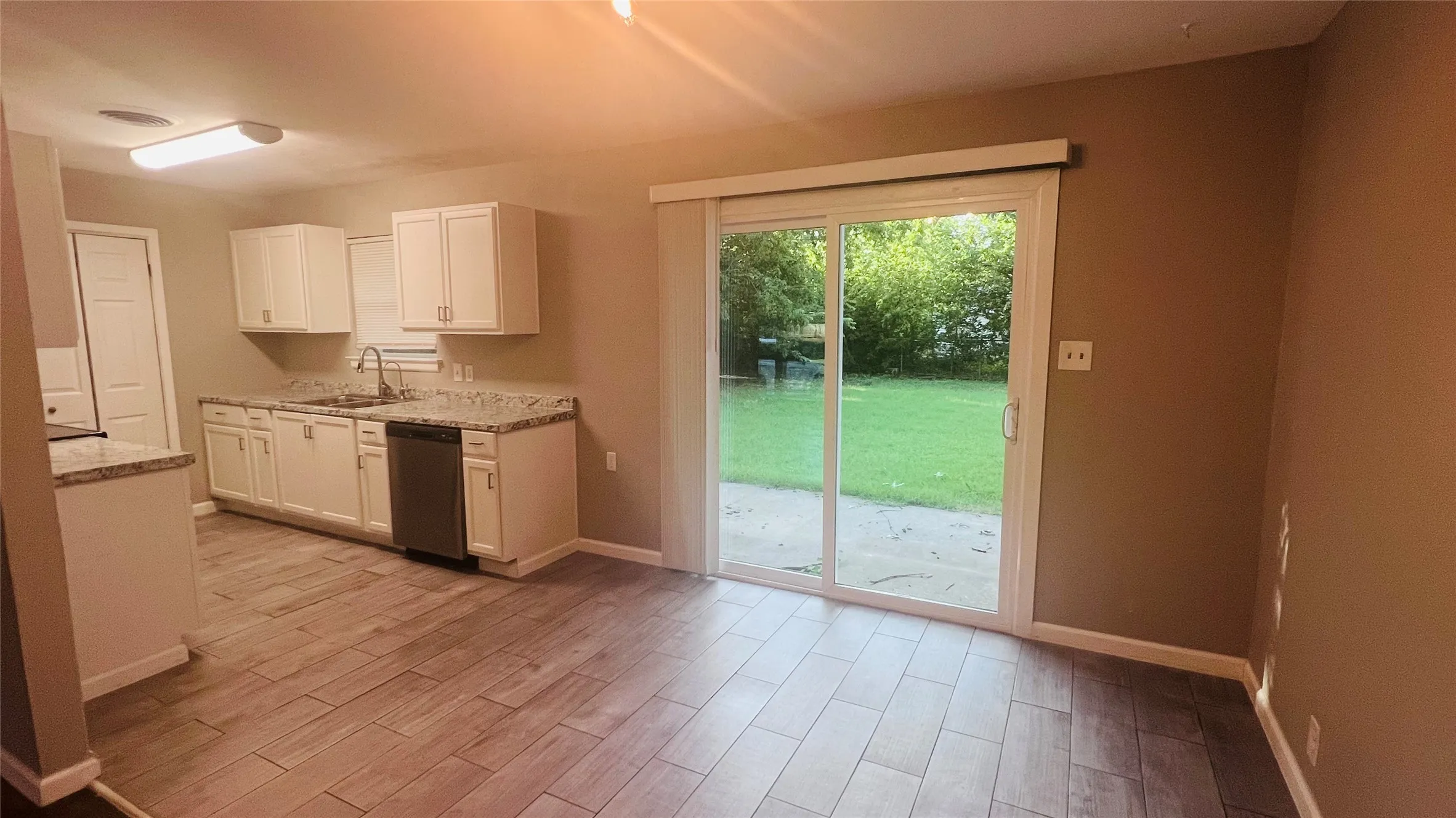 Open-concept kitchen and dining area featuring wood-finish flooring, white cabinetry, a stainless steel dishwasher, and a sliding glass door leading to a patio and grassy yard
