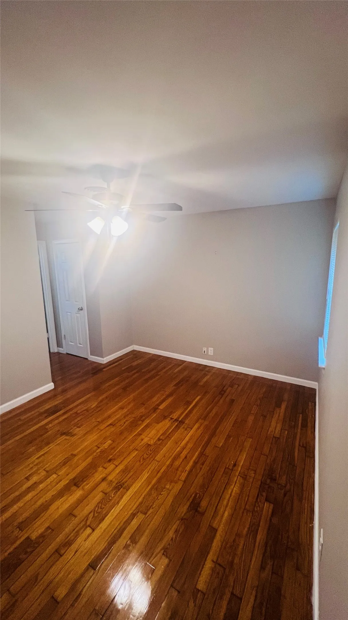 Room featuring rich wood-finish flooring, light gray walls, white baseboards, a ceiling fan with integrated lighting, and a white interior door
