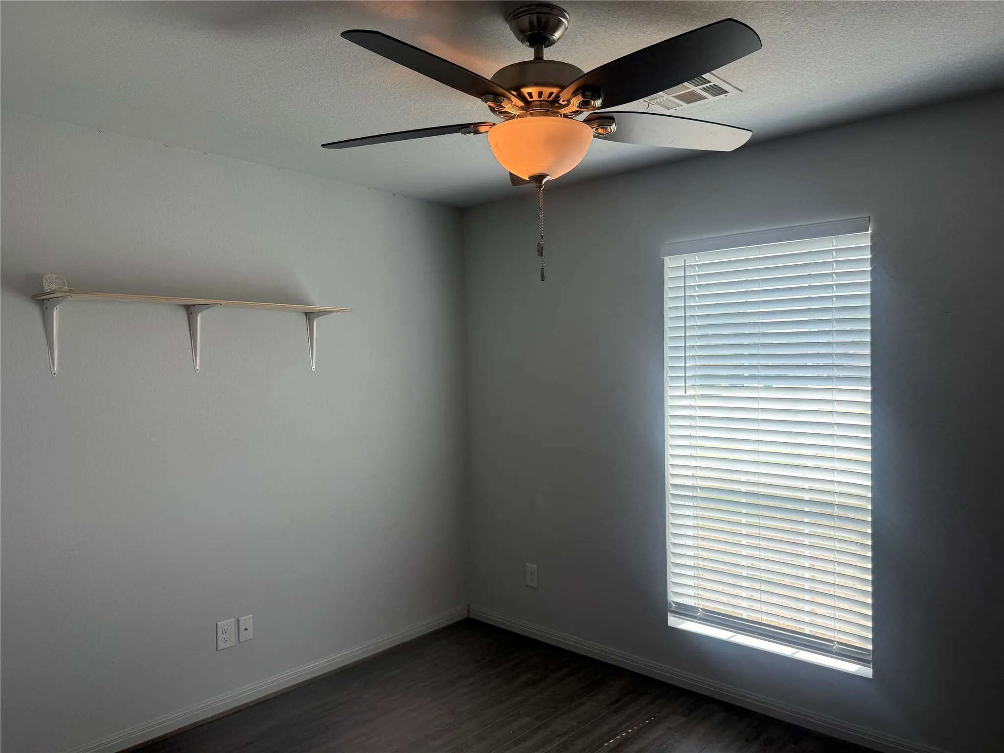 Dark wood-finish flooring with light gray walls, featuring a window with horizontal blinds and a ceiling fan with integrated lighting