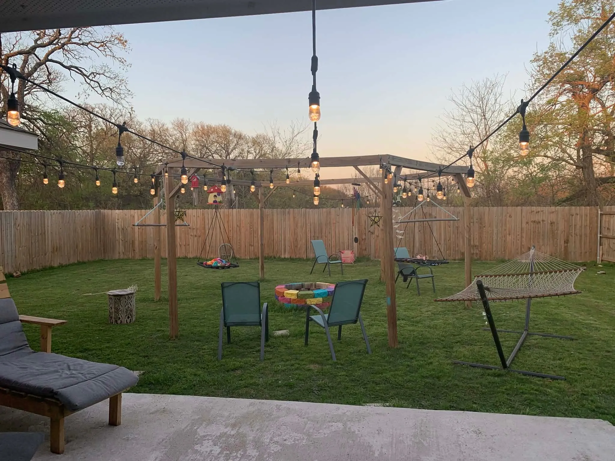 Expansive backyard featuring a privacy fence, string lighting, a wooden pergola structure, a concrete patio area, and a large grassy lawn