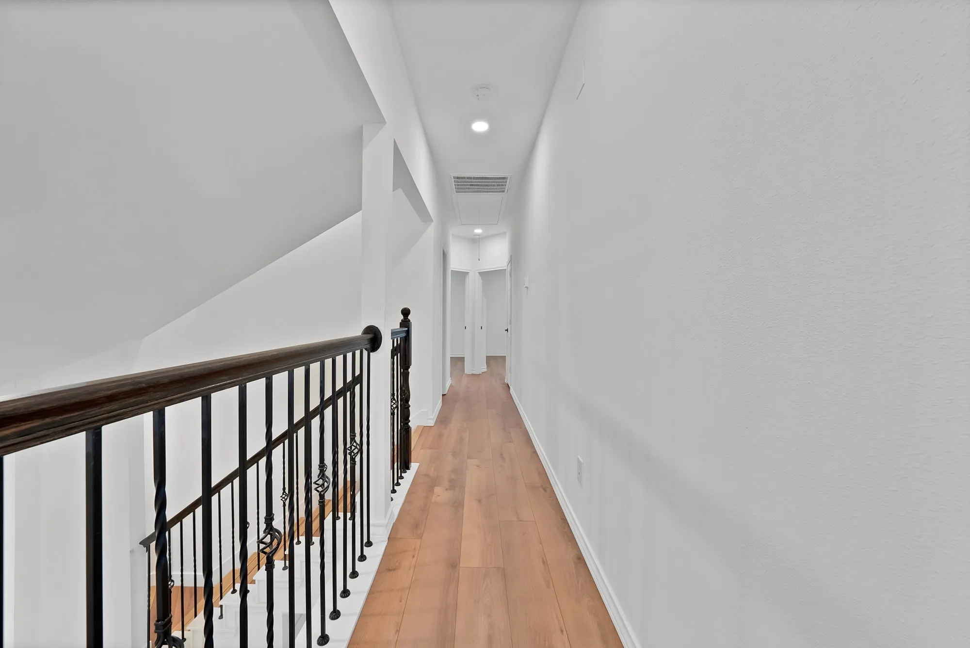 Upper-level hallway featuring wood-finish flooring, recessed lighting, and a wrought iron baluster railing with a dark wood handrail