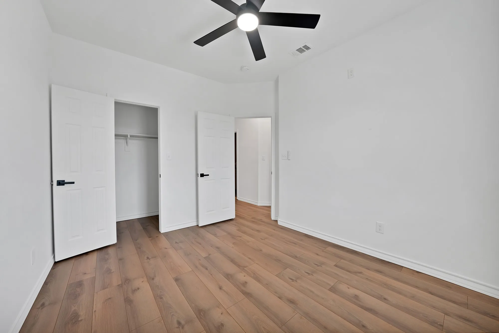 Spacious interior room featuring wood-finish flooring, white walls, and a ceiling fan with black blades