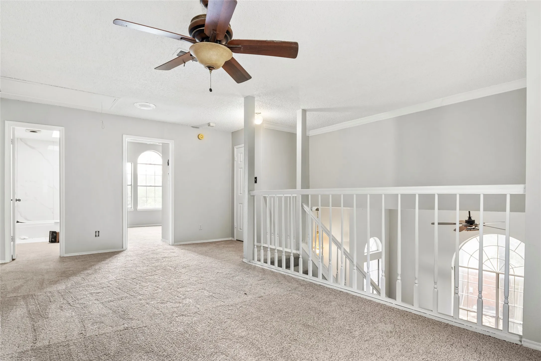 Carpeted upper-level landing featuring a wood-tone ceiling fan, white railing overlooking the lower level, arched windows, and white trim molding