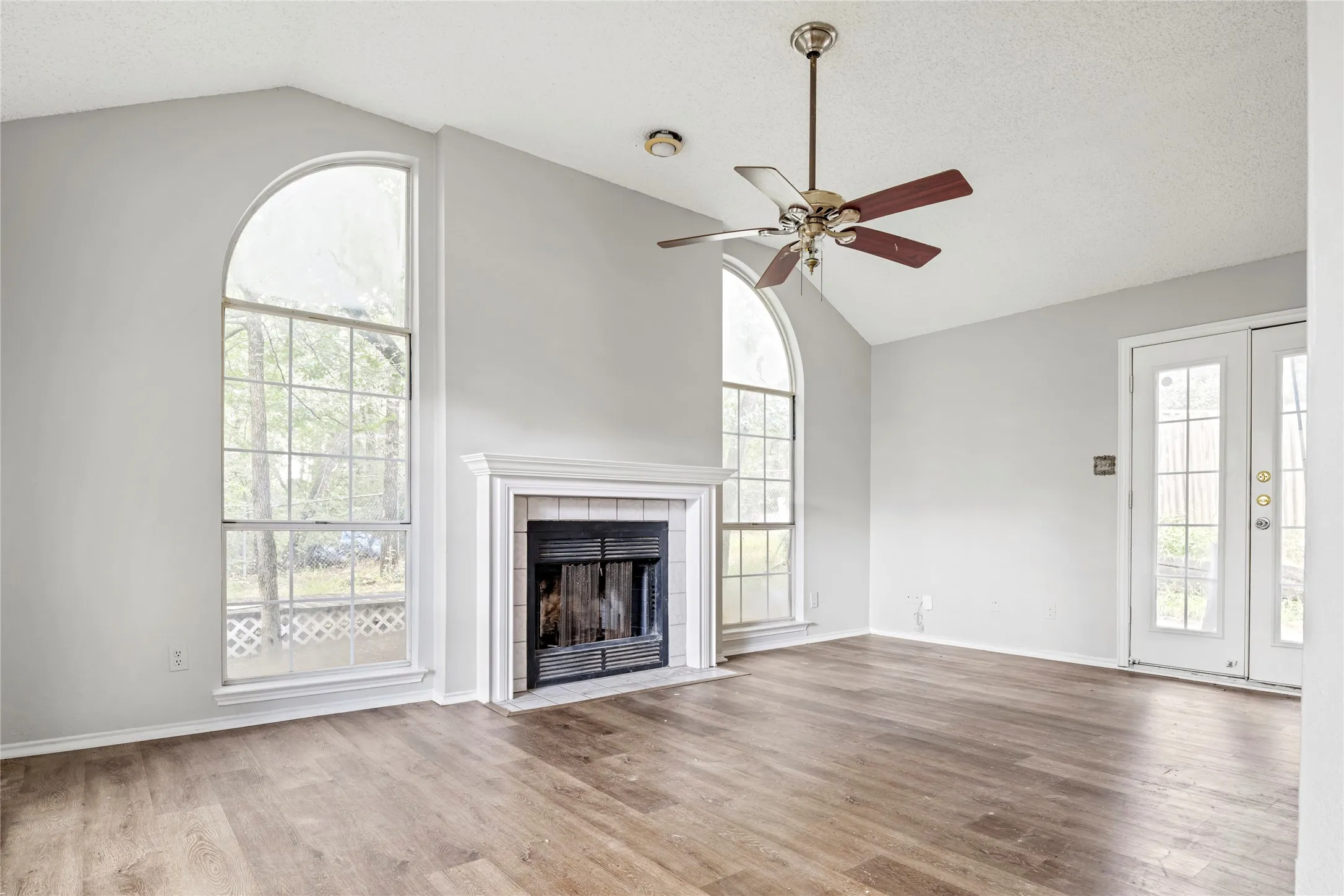 Vaulted ceilings with a ceiling fan, fireplace with a white mantel, two arched windows, wood-finish flooring, and French doors