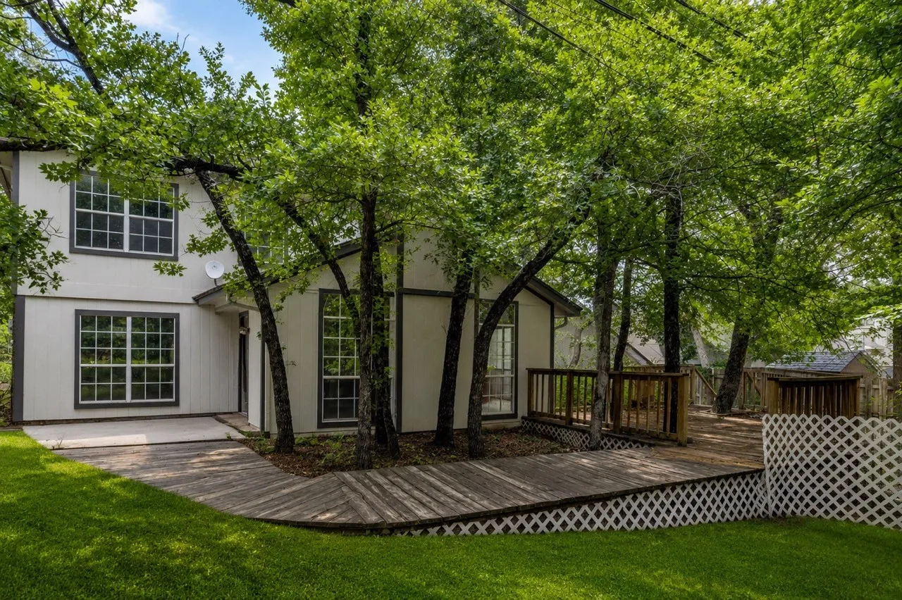 Exterior featuring white siding with dark trim, multiple large windows with grid patterns, a wooden deck, mature trees, and a manicured lawn