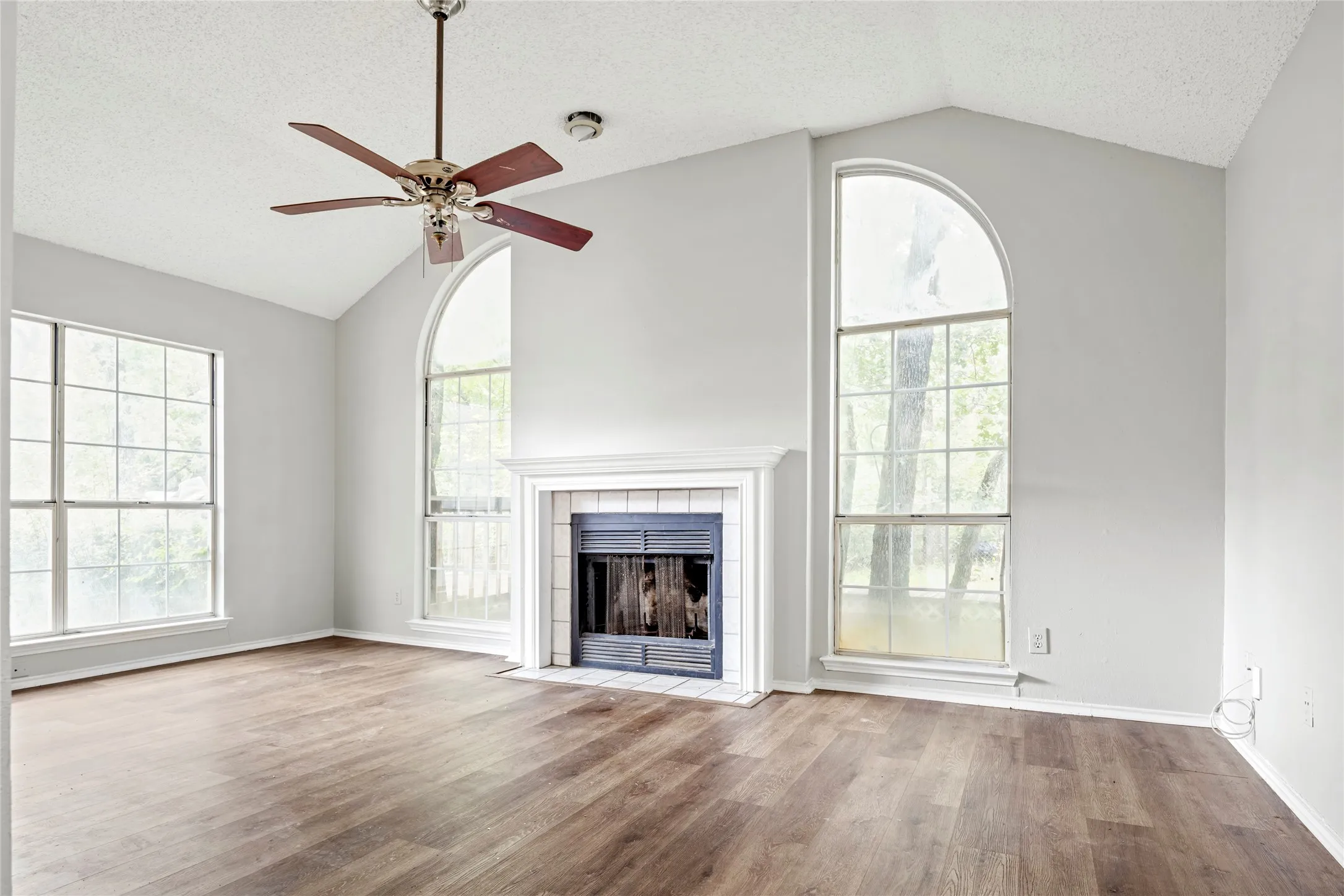 Living area featuring vaulted ceilings, wood-finish flooring, a fireplace with a white mantel, and multiple windows including a prominent arched window