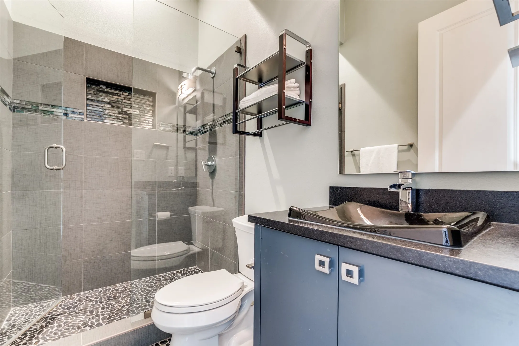 Bathroom featuring a glass-enclosed shower with pebble flooring and a tiled accent niche, a toilet, a vanity with a black countertop, a vessel sink, and a wall-mounted mirror