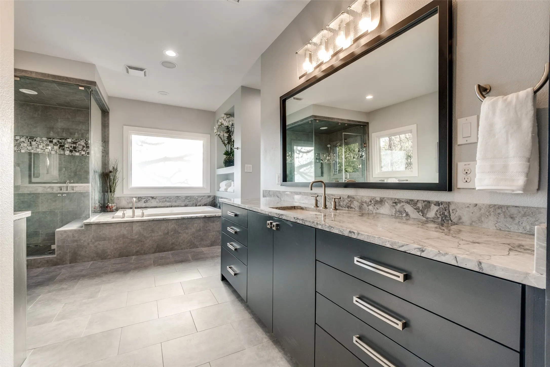 Large bathroom featuring a dual vanity with a light-toned countertop, a large framed mirror, a built-in soaking tub, a separate glass-enclosed shower, and light gray tile flooring