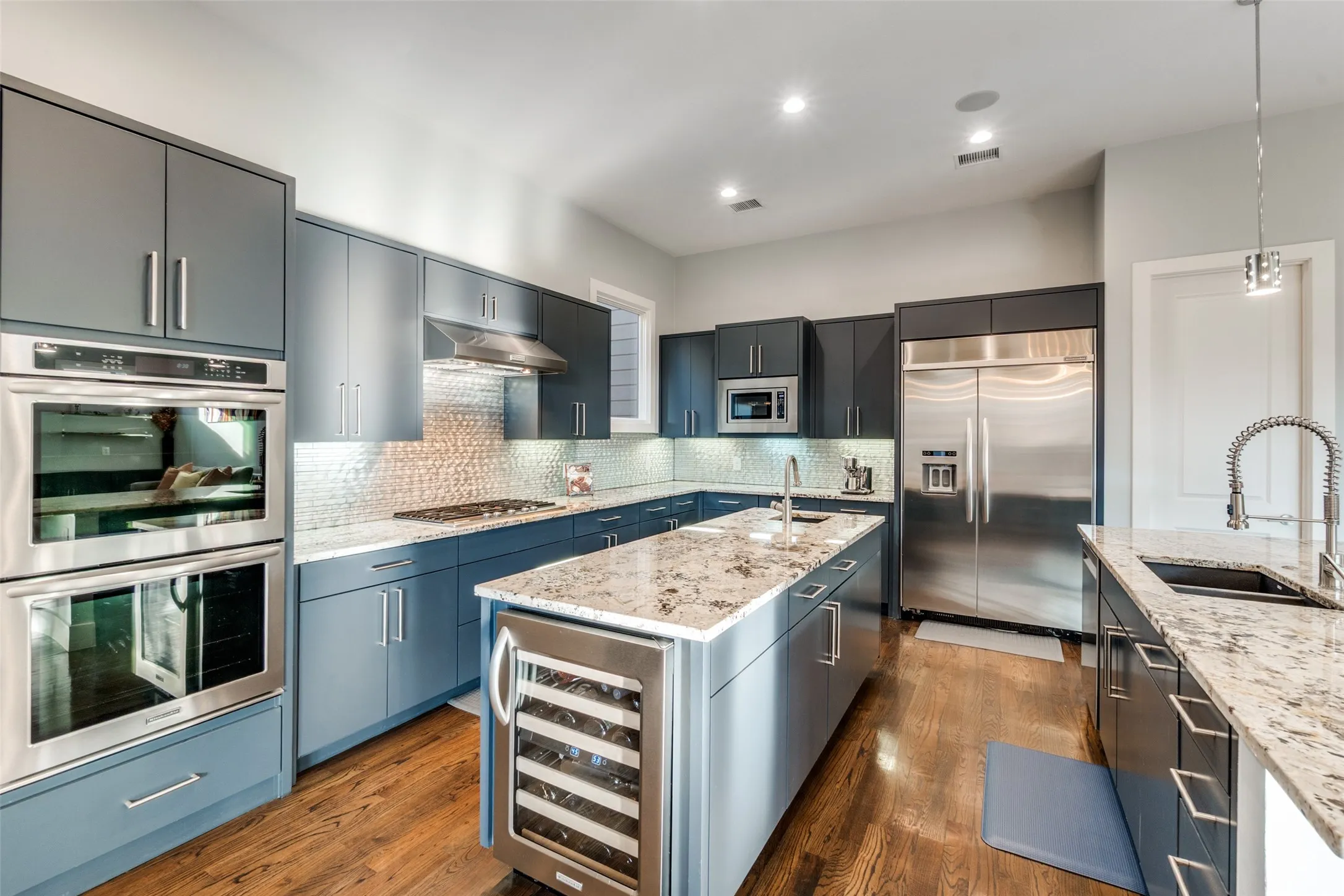 Modern kitchen featuring wood-finish flooring, blue cabinetry, light granite countertops, stainless steel appliances, and a mosaic tile backsplash