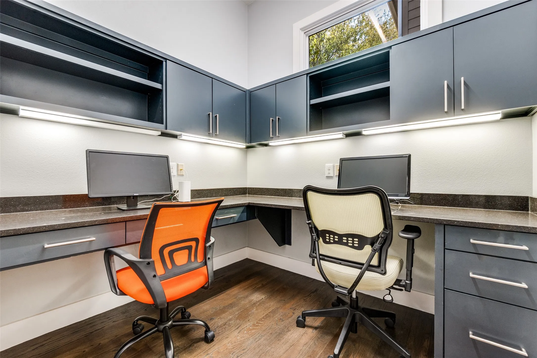 Built-in corner desk area featuring dark wood-finish flooring, ample cabinetry with silver hardware, under-cabinet task lighting, and a high window