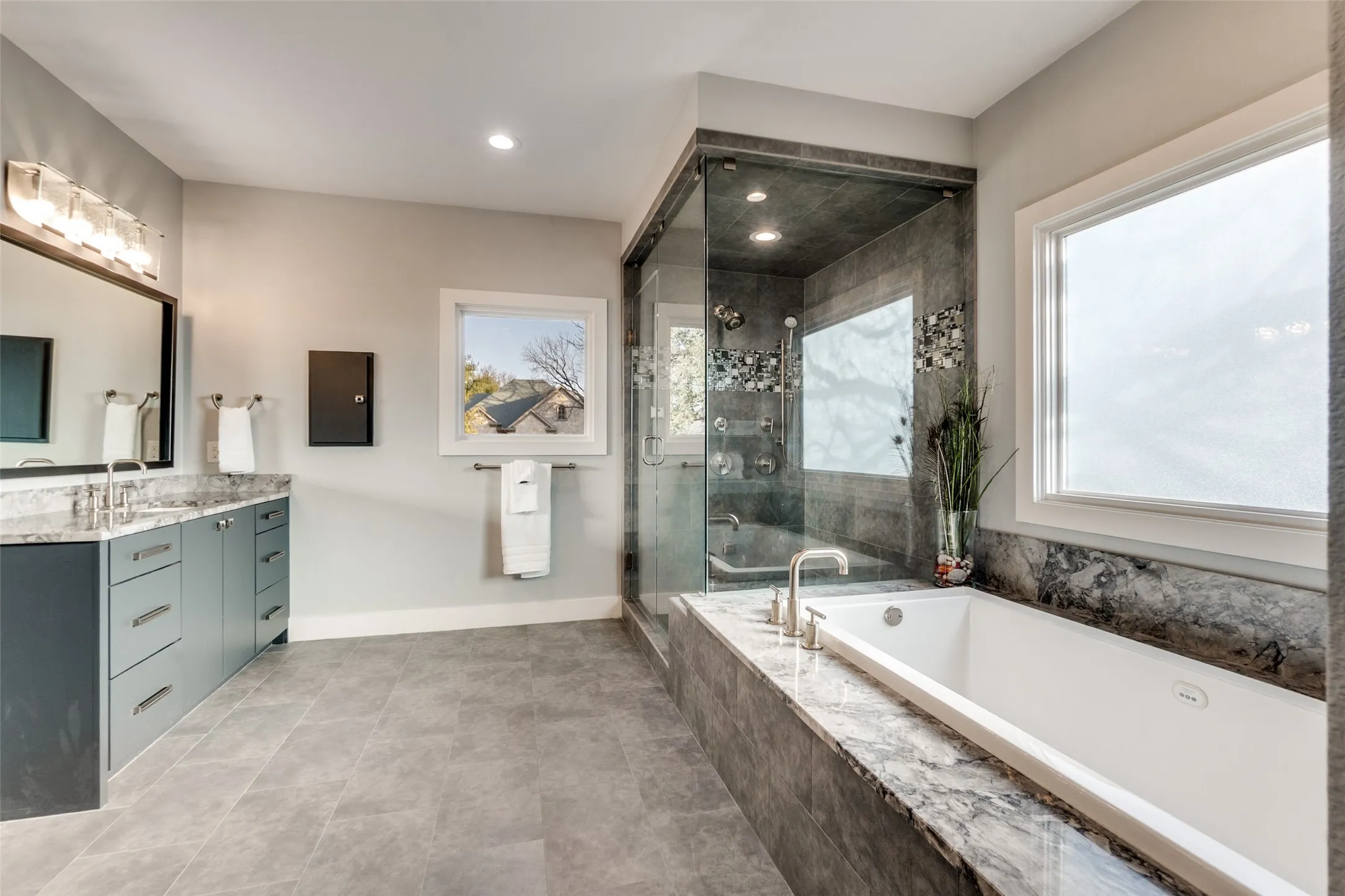 Luxurious bathroom featuring a built-in soaking tub, a large walk-in glass shower with stone tile, a double vanity with stone countertops, and gray tile flooring