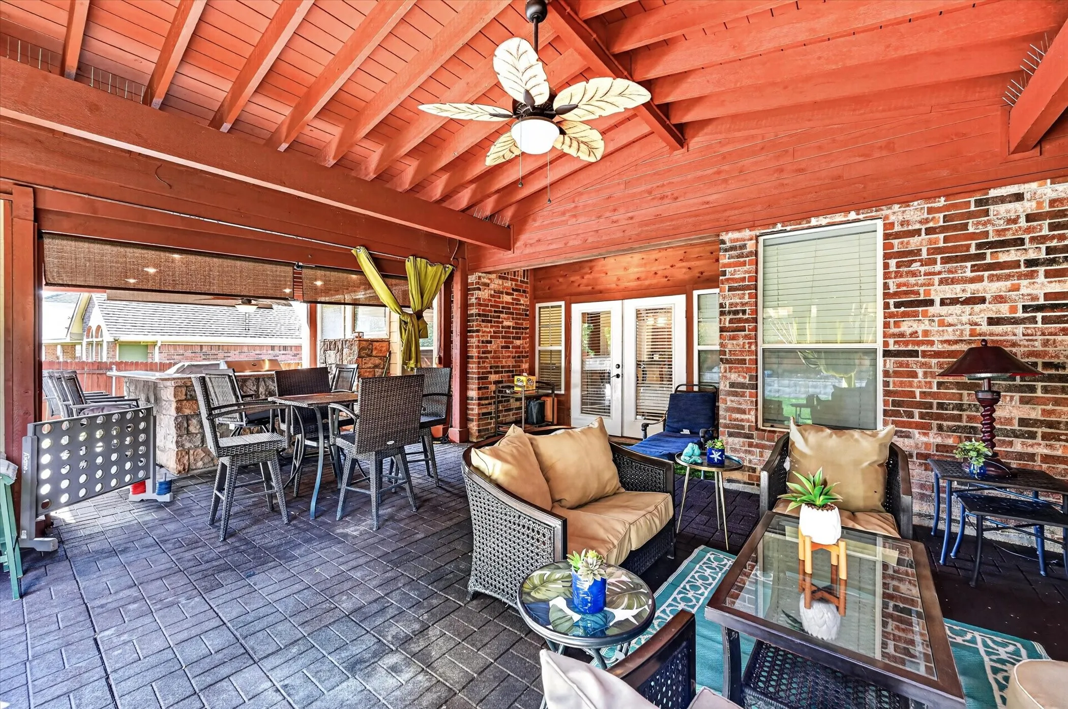 Expansive covered patio featuring a paver stone floor, exposed wood beam ceiling, ceiling fan, and a built-in outdoor kitchen with stone veneer