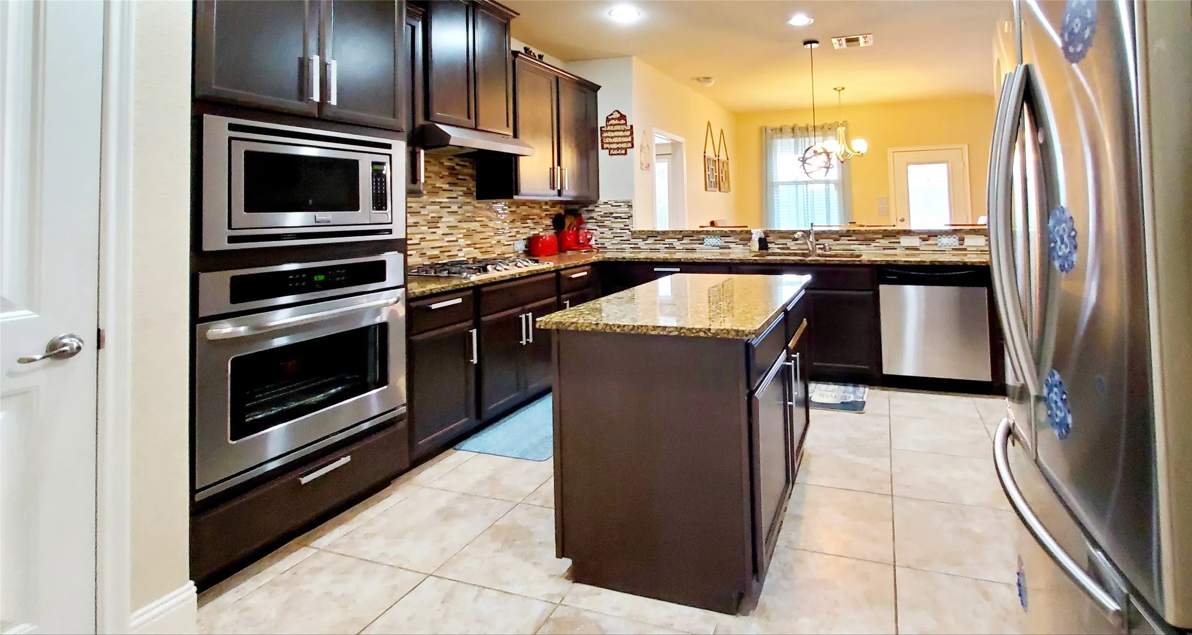 Kitchen featuring dark wood cabinetry, granite countertops, an integrated stainless steel oven and microwave combination, a mosaic tile backsplash, and a central island with additional storage
