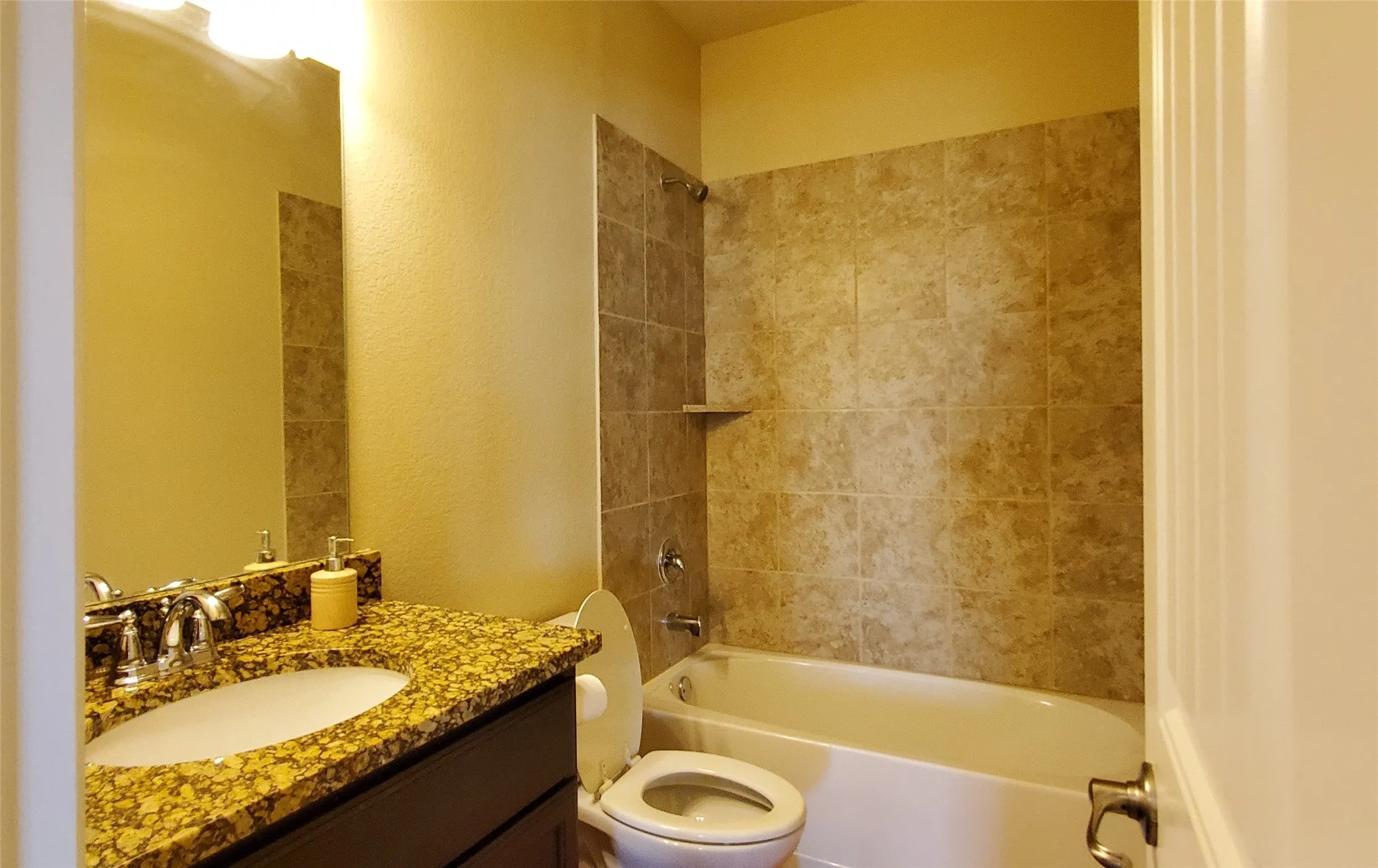 Bathroom featuring a white bathtub, ceramic tile shower surround with integrated shelf, vanity with a granite countertop, undermount sink, and chrome fixtures