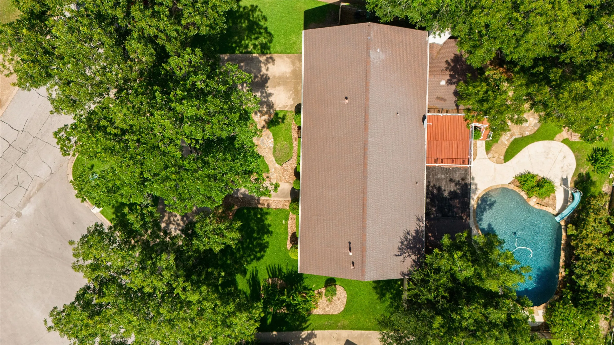 Aerial perspective highlighting a reddish shingle roof, lush green lawn, mature well maintained pecan trees in the front and back yards, and a freeform swimming pool with an attached slide, a flagstone patio area with an inground gas conection for a gas cooker or a future outdoor kitchen