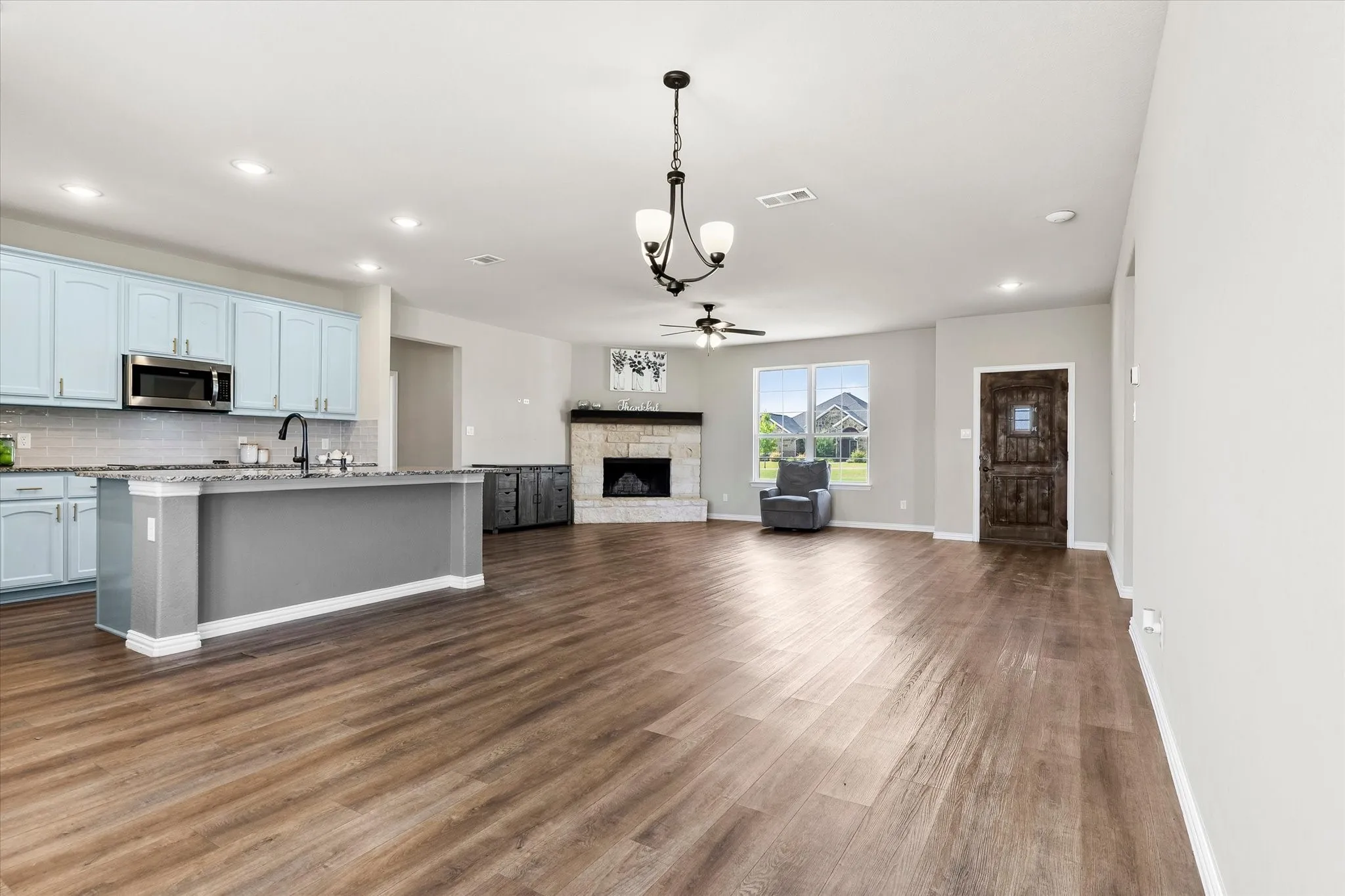 Spacious open-concept interior featuring wood-finish flooring, a kitchen island with a dark countertop, light blue cabinetry with stainless steel appliances, a stone-front fireplace, and recessed lighting