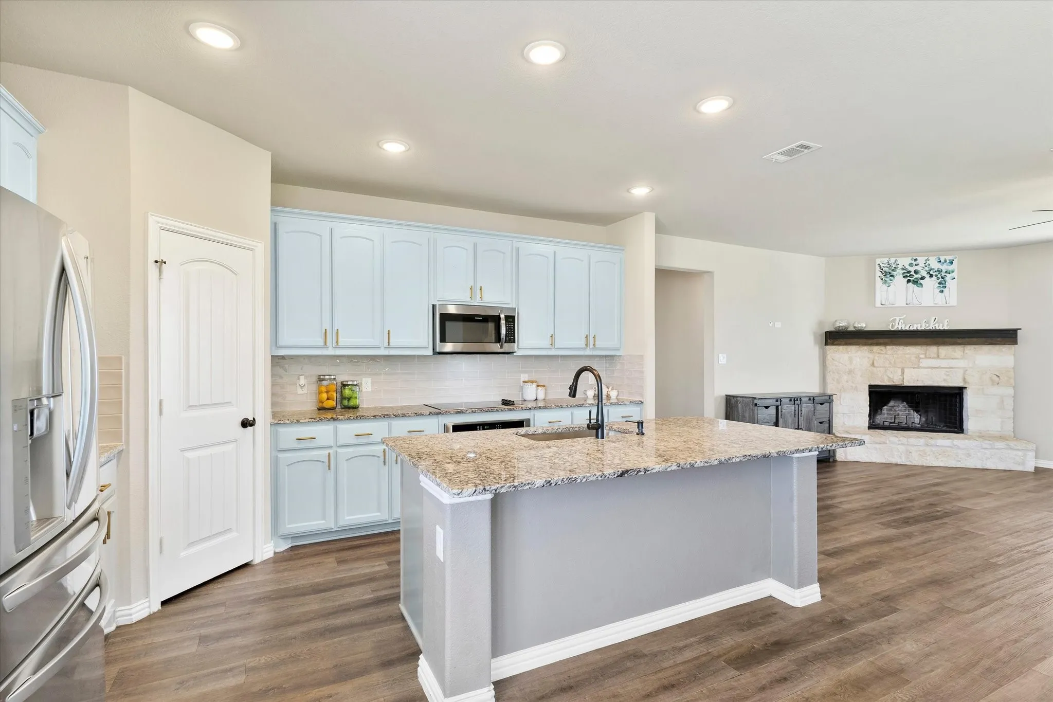 Open-concept kitchen and living area featuring white cabinetry, stainless steel appliances, granite-style countertops, a large center island with seating, wood-finish flooring throughout, recessed lighting, and a fireplace with stone surround visible in the adjoining space