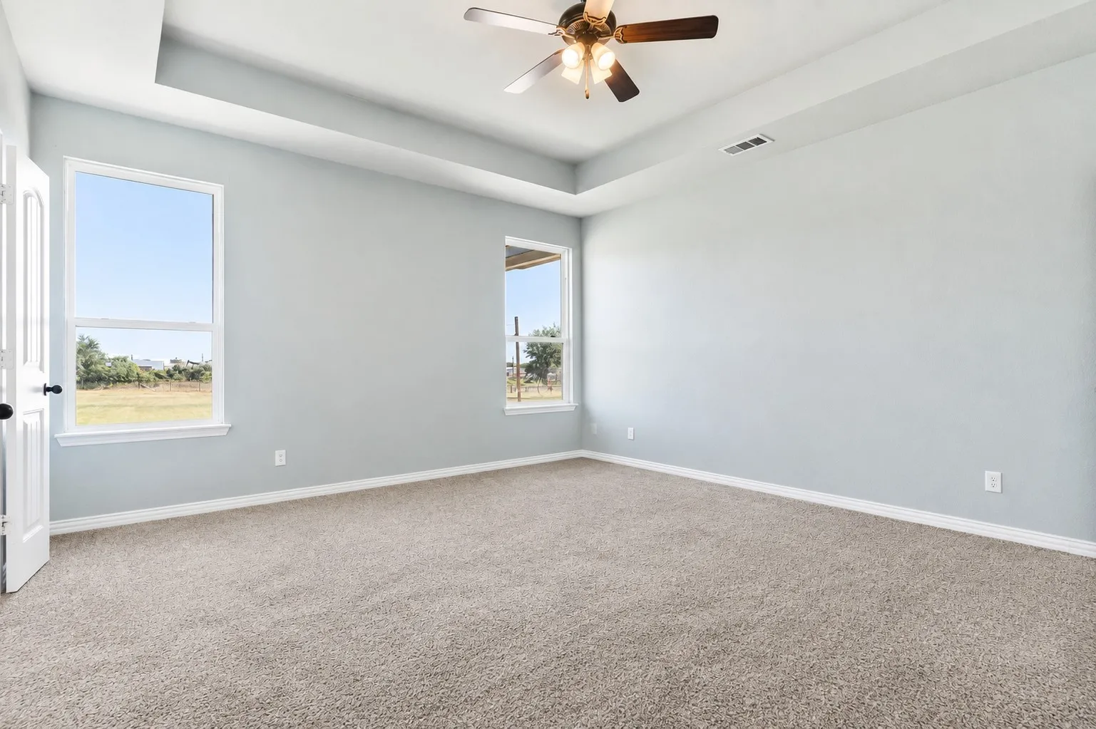 Spacious bedroom showcasing neutral wall finishes, plush carpet flooring, recessed ceiling detail with ceiling fan, and abundant natural light from dual windows with expansive outdoor vistas