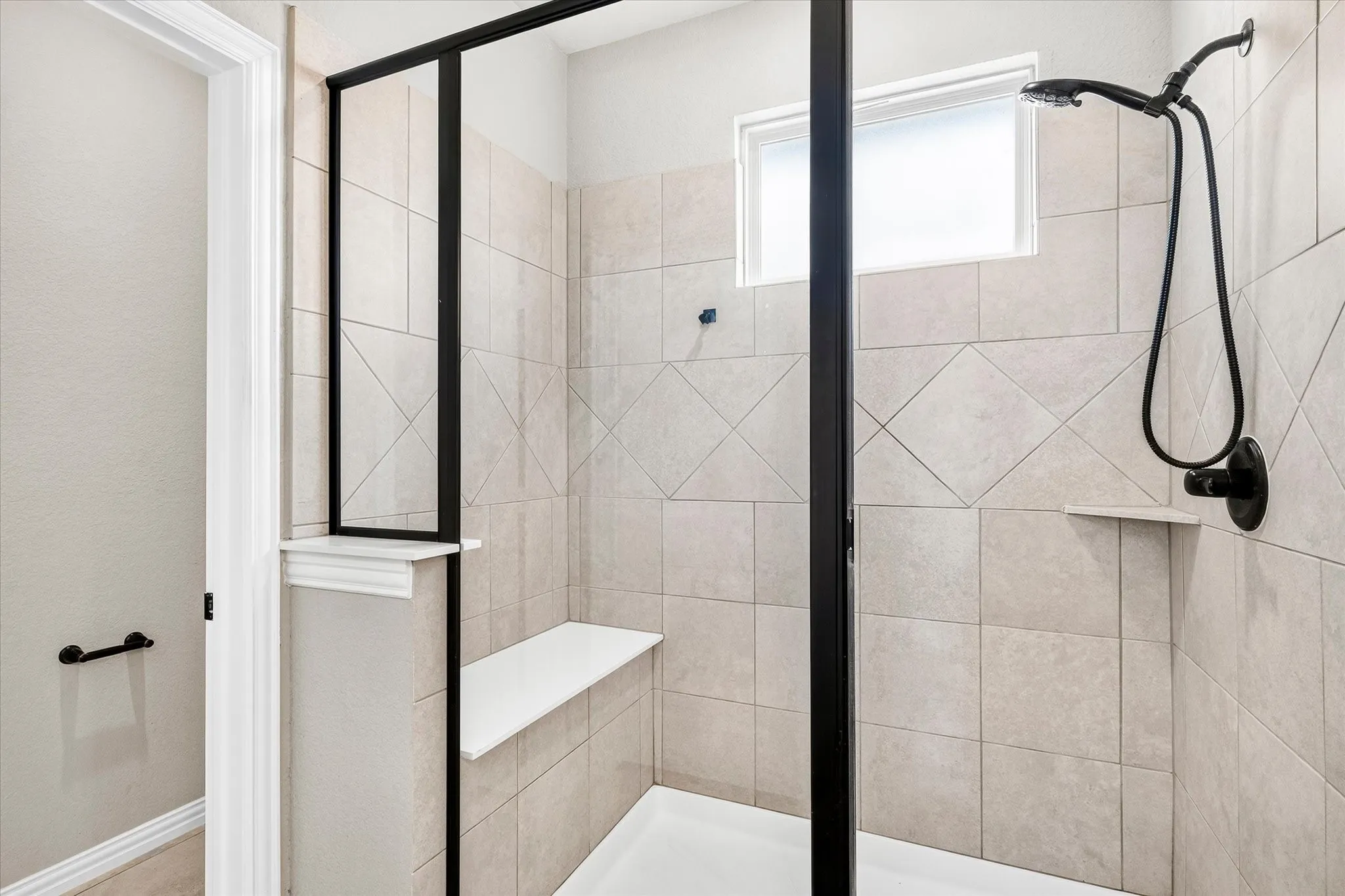 Modern primary bathroom featuring a frameless glass shower enclosure with matte black hardware, neutral tile walls laid in diagonal pattern, built-in bench seating, and contemporary rainfall showerhead with handheld spray fixture