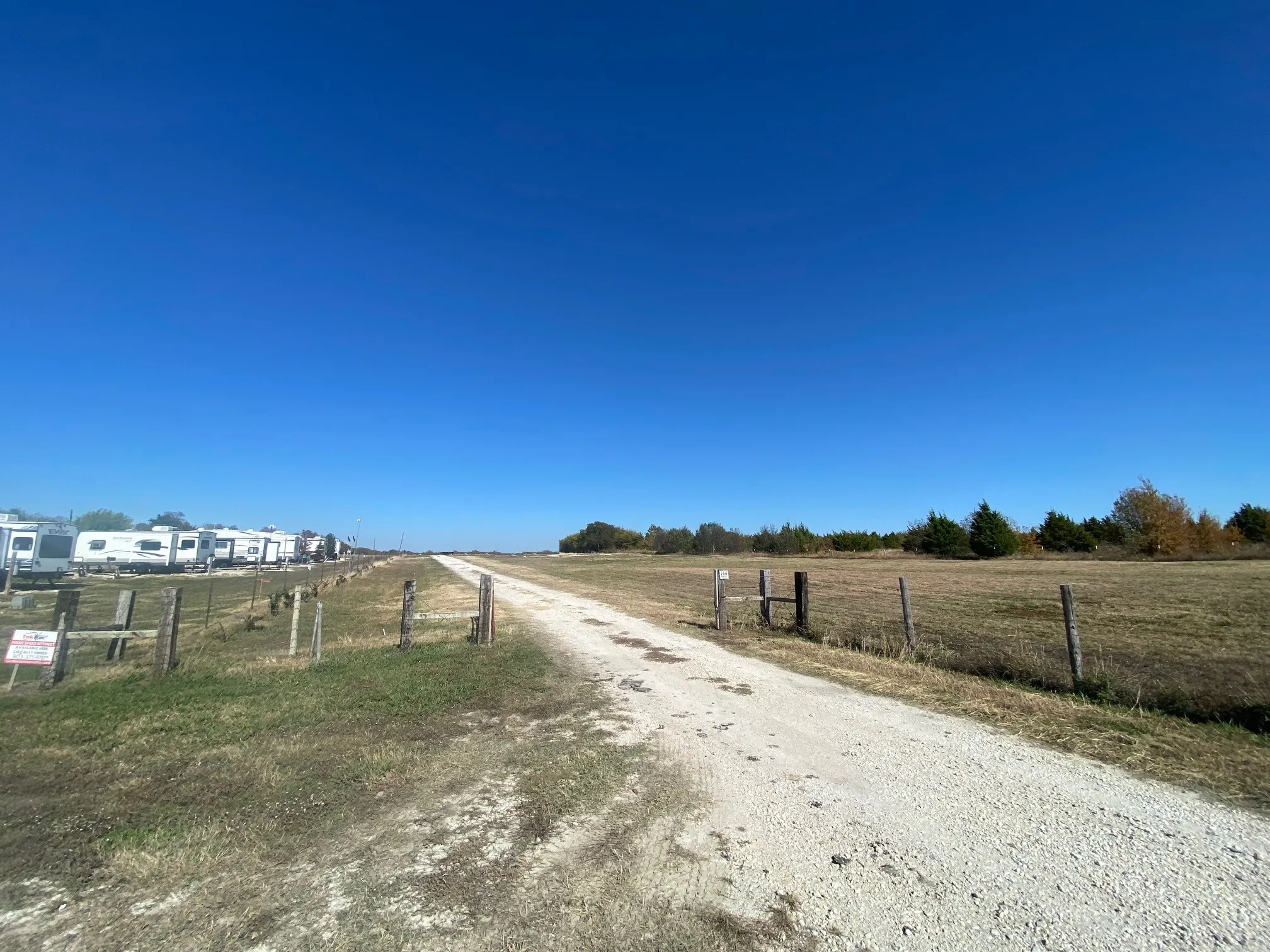 Gravel road extending through open land with a wooden post fence line and distant trees