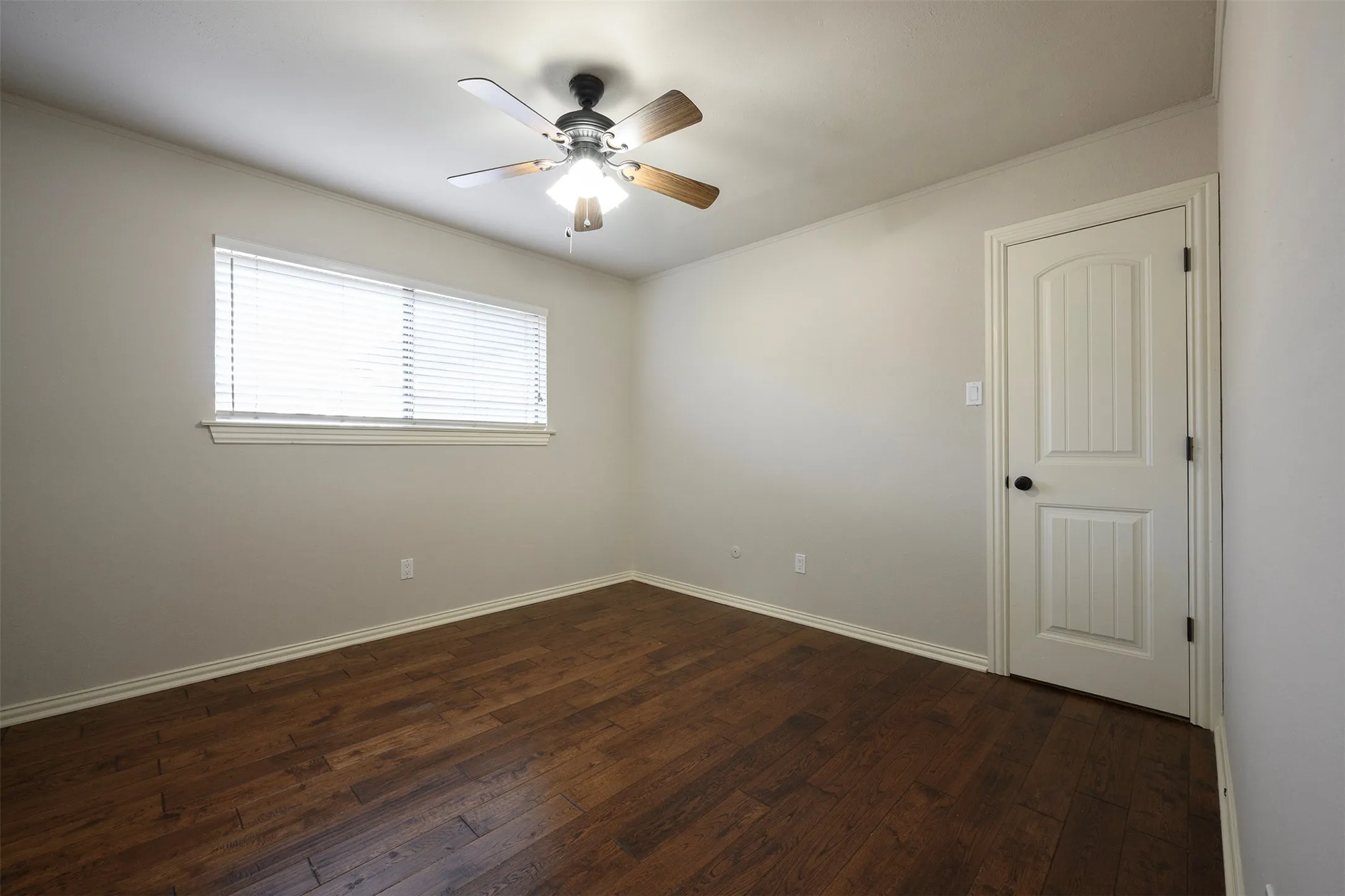 Spacious room featuring wood-finish flooring, a window with horizontal blinds, a ceiling fan with integrated lighting, a paneled door with a matte black knob, and neutral wall paint