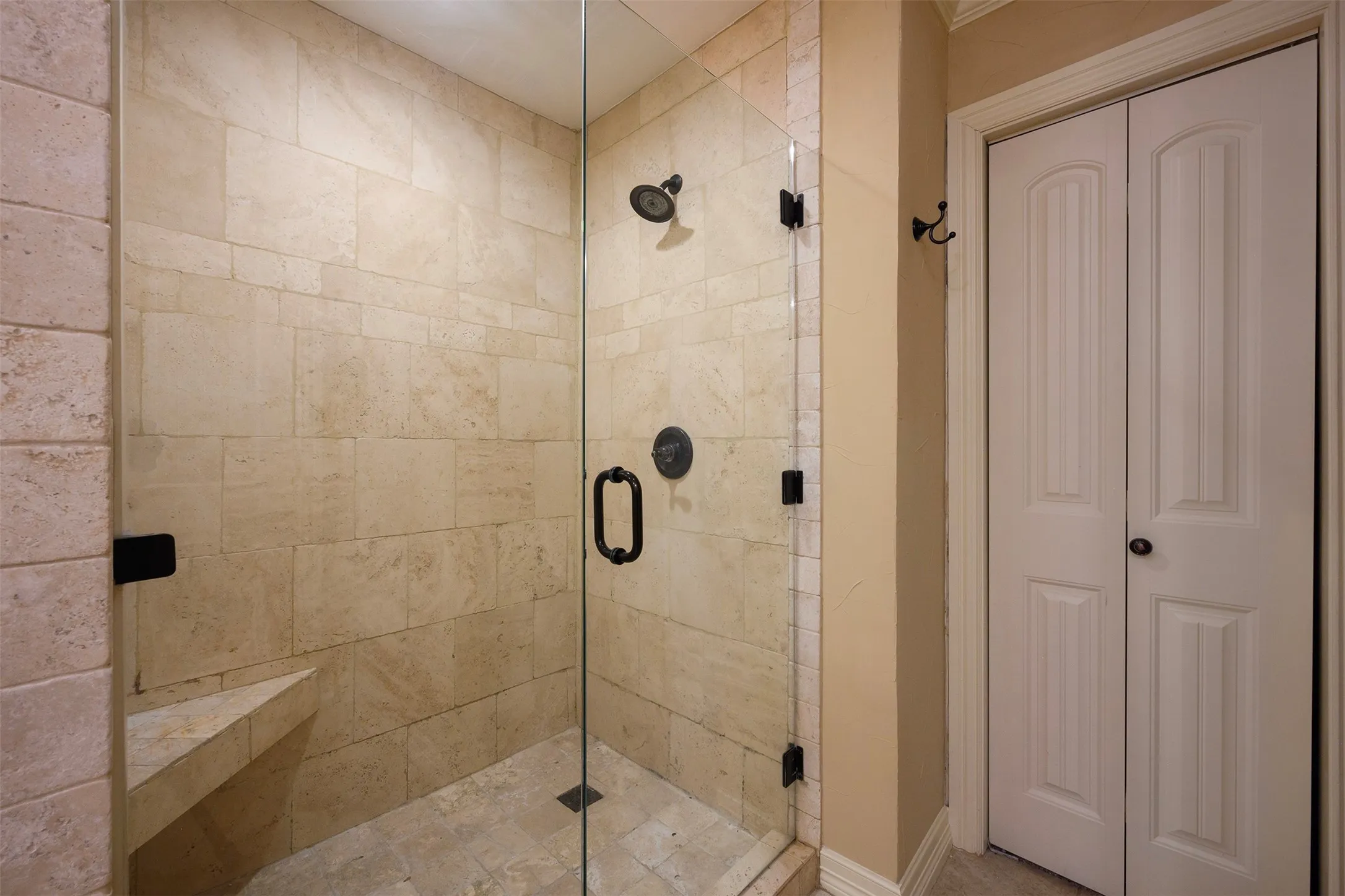 Glass-enclosed shower with stone tile walls and flooring, featuring a built-in stone bench and matte black fixtures