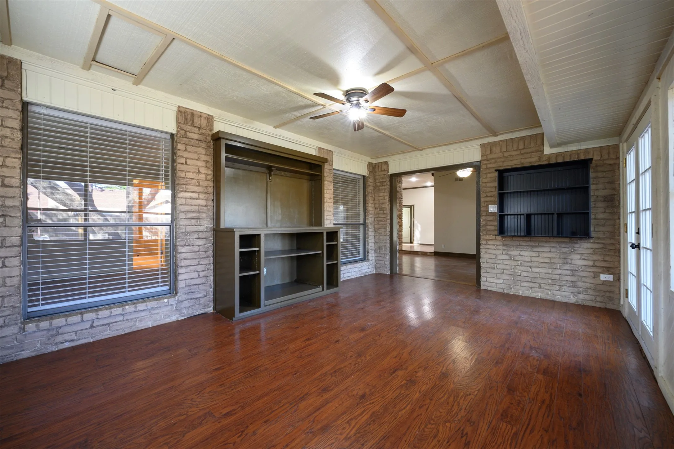 Spacious room featuring wood-finish flooring, exposed brick walls, a built-in entertainment center, and a ceiling fan