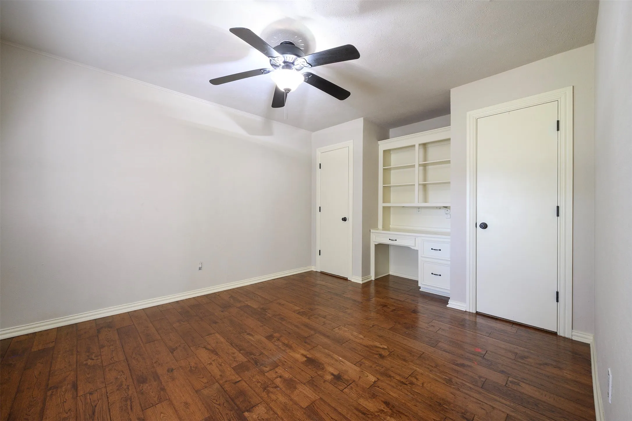 Wood-finish flooring throughout, featuring a built-in desk with shelving, painted white walls, white baseboards, and a ceiling fan with integrated lighting