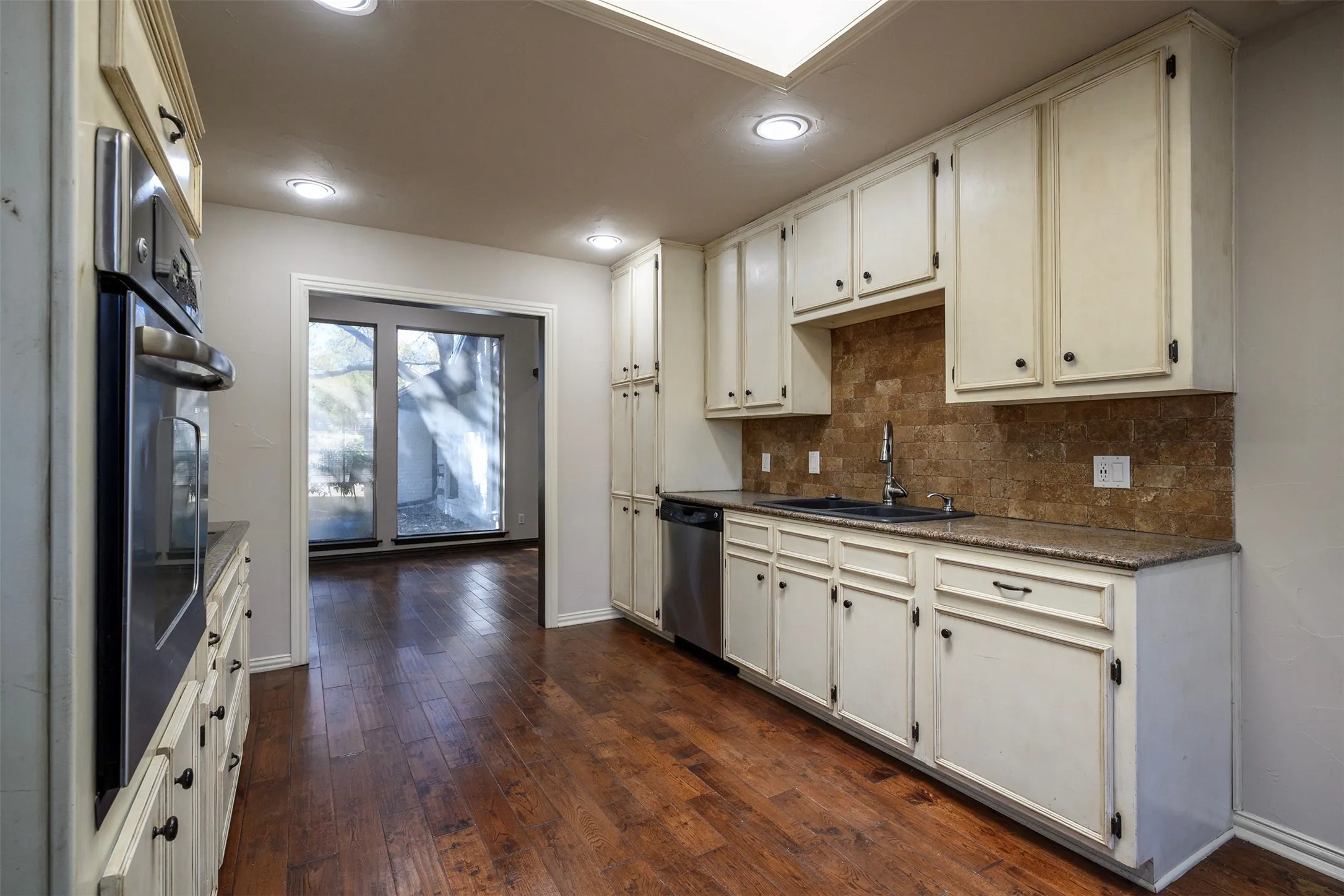 Kitchen featuring wood-finish flooring, cream-toned cabinetry, stainless steel dishwasher, built-in oven, and a tiled backsplash