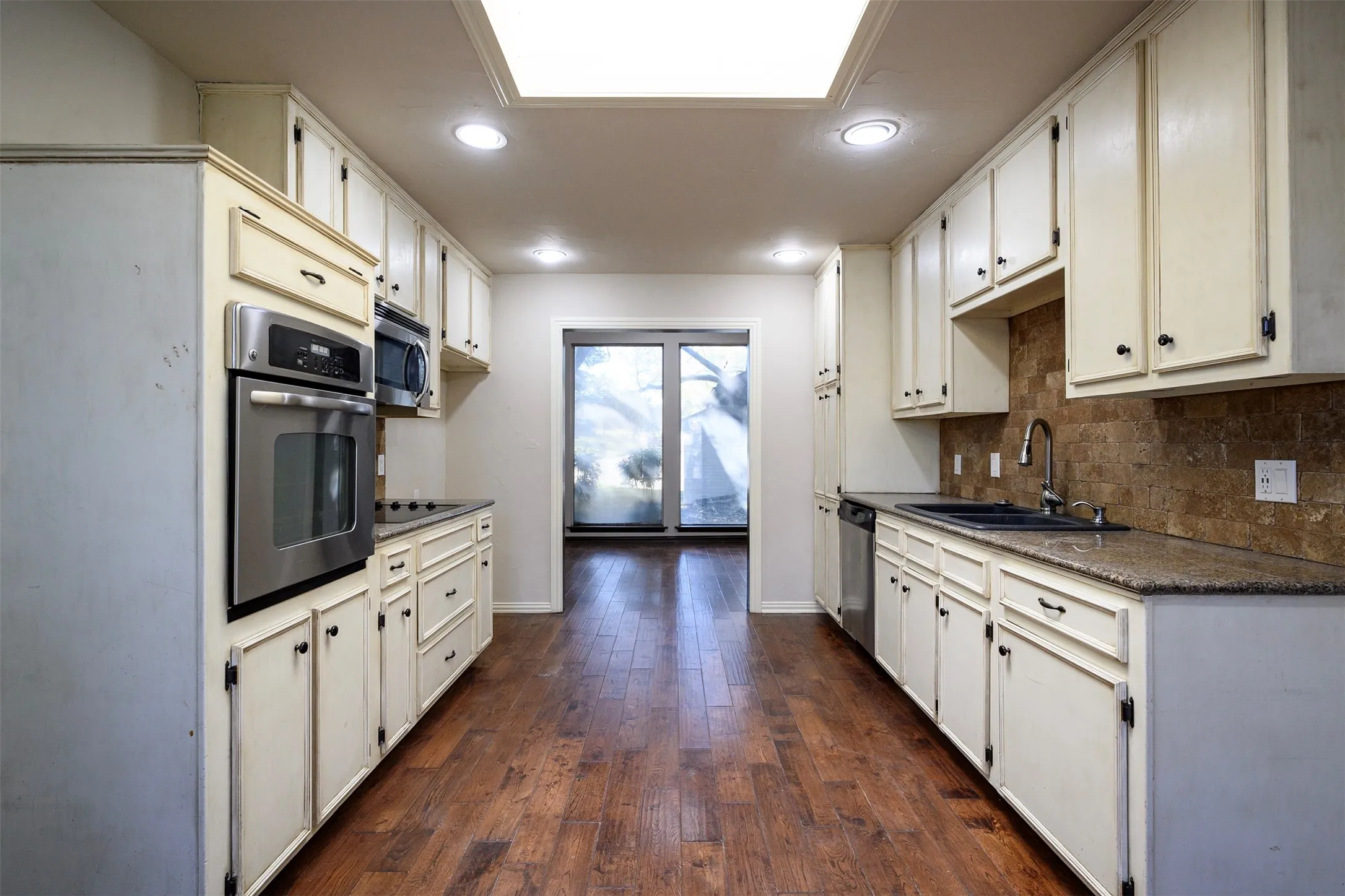 Galley kitchen featuring wood-finish flooring, white cabinetry, integrated stainless steel appliances, speckled countertops, and a stone-tiled backsplash