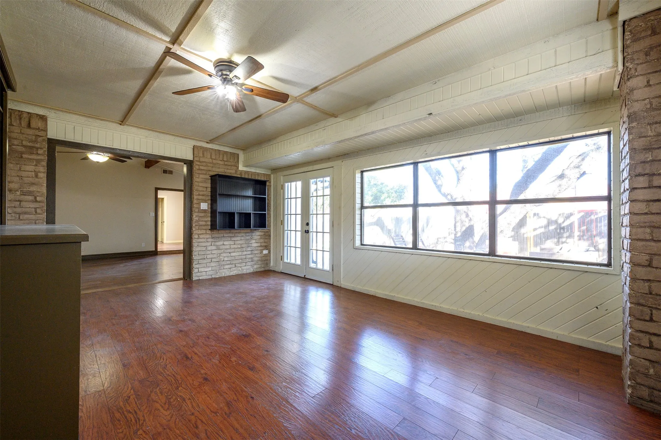 Spacious room with wood-finish flooring, exposed beam ceiling with ceiling fan, large picture windows, and brick architectural accents