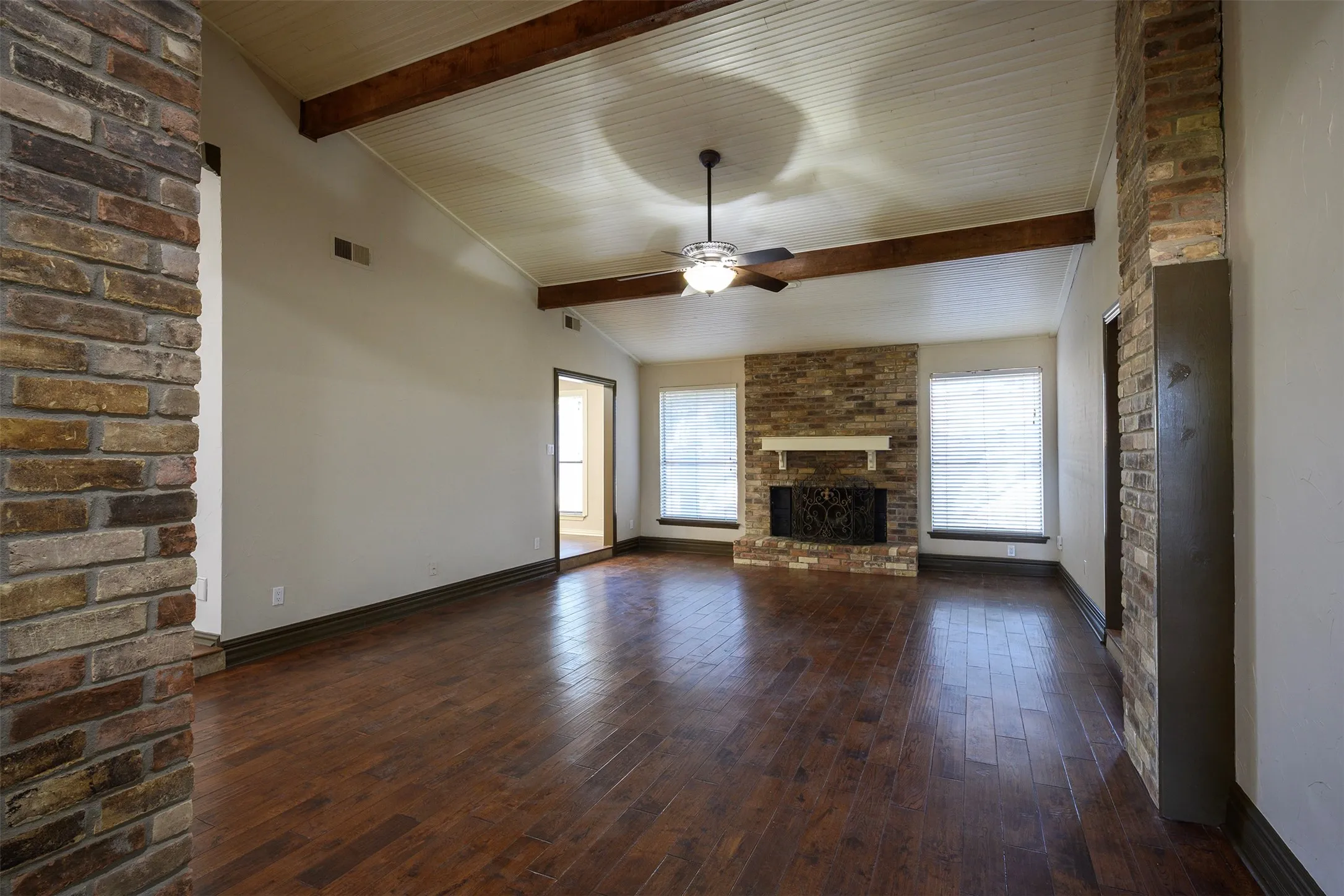 Expansive room featuring wood-finish flooring, a vaulted ceiling with exposed beams, and a brick fireplace with a white mantel