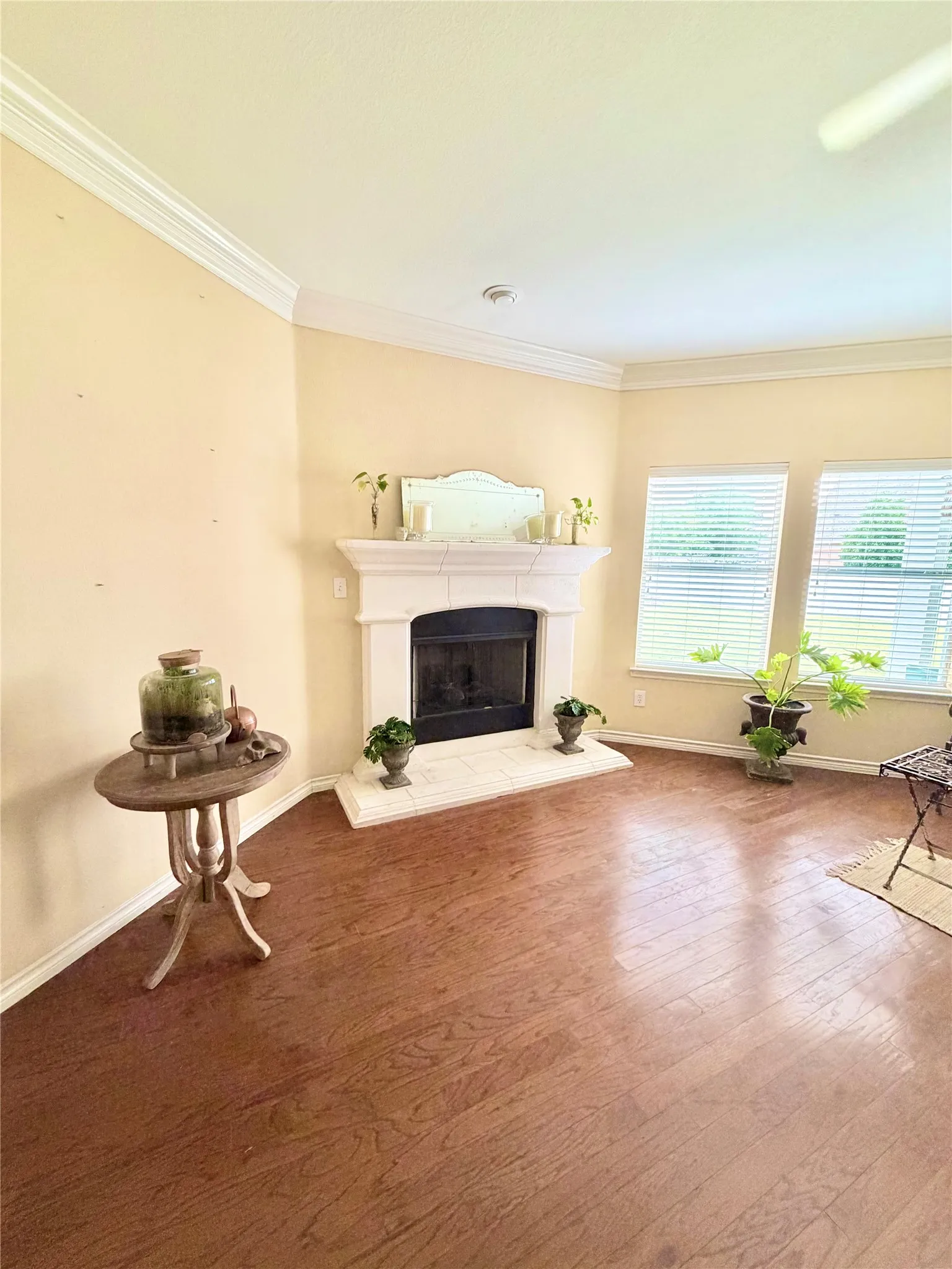 Living area featuring a stone fireplace with hearth, wood-finish flooring, two large windows with blinds, and detailed crown molding