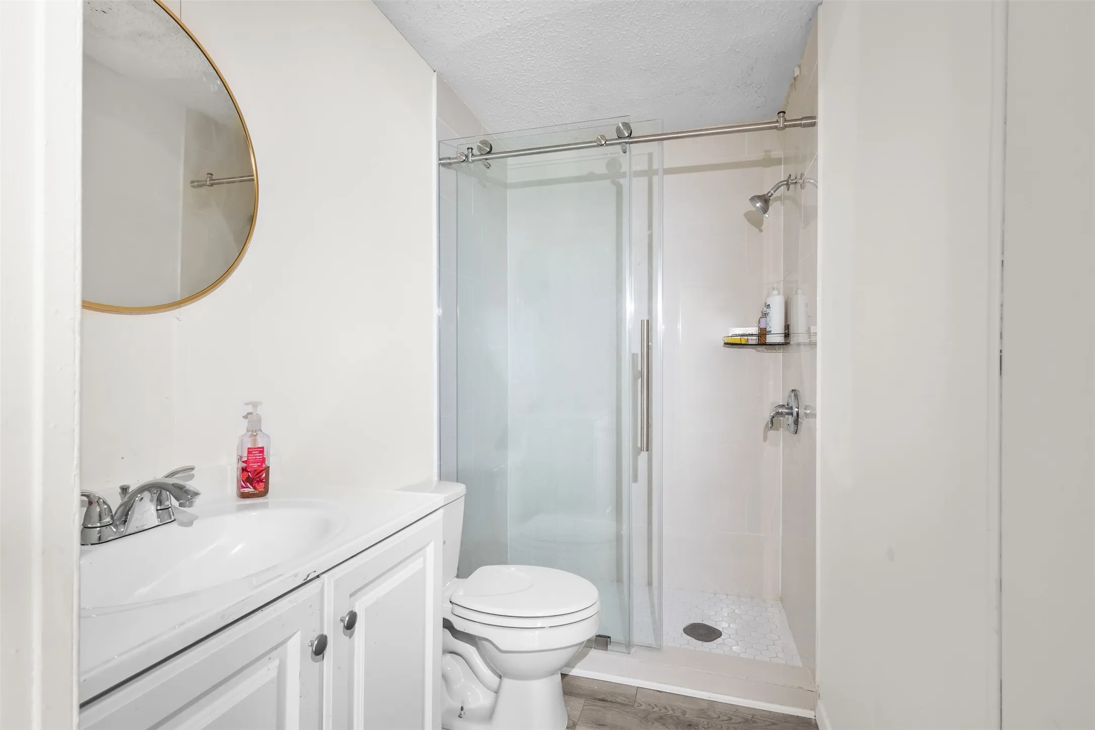 Bathroom featuring a white vanity with an integrated sink, a circular mirror with a gold-tone frame, a toilet, and a glass-enclosed shower with a tiled floor