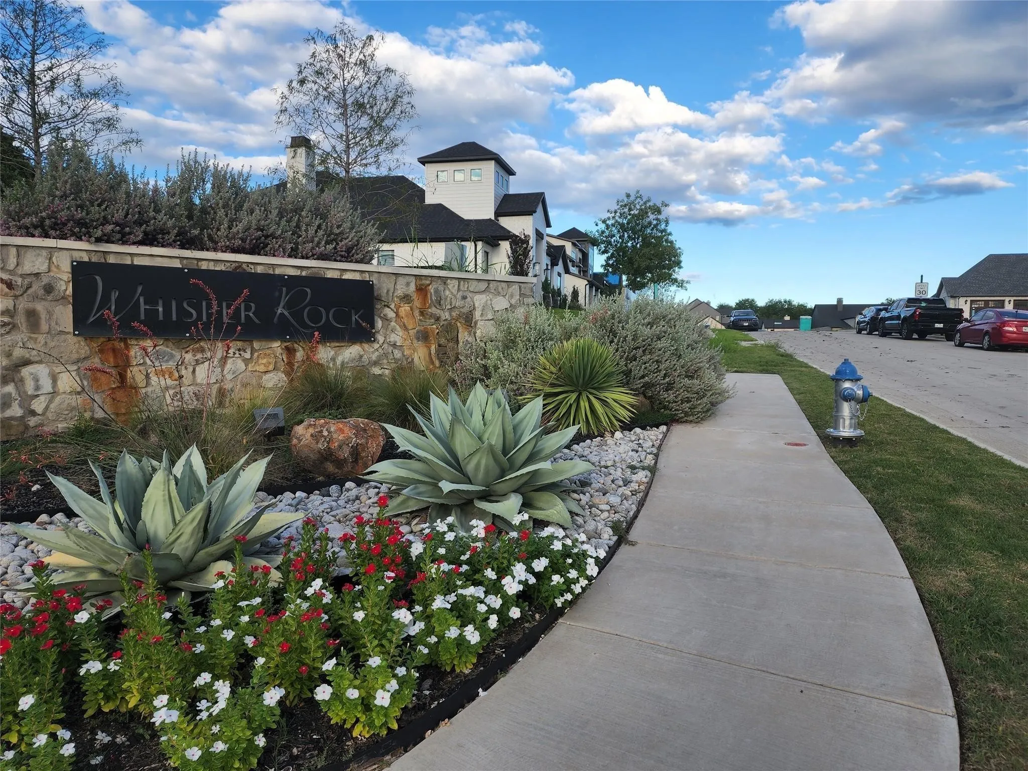 Stone entry monument with integrated sign, lush landscaping featuring agaves and flowering plants, and a concrete pedestrian path