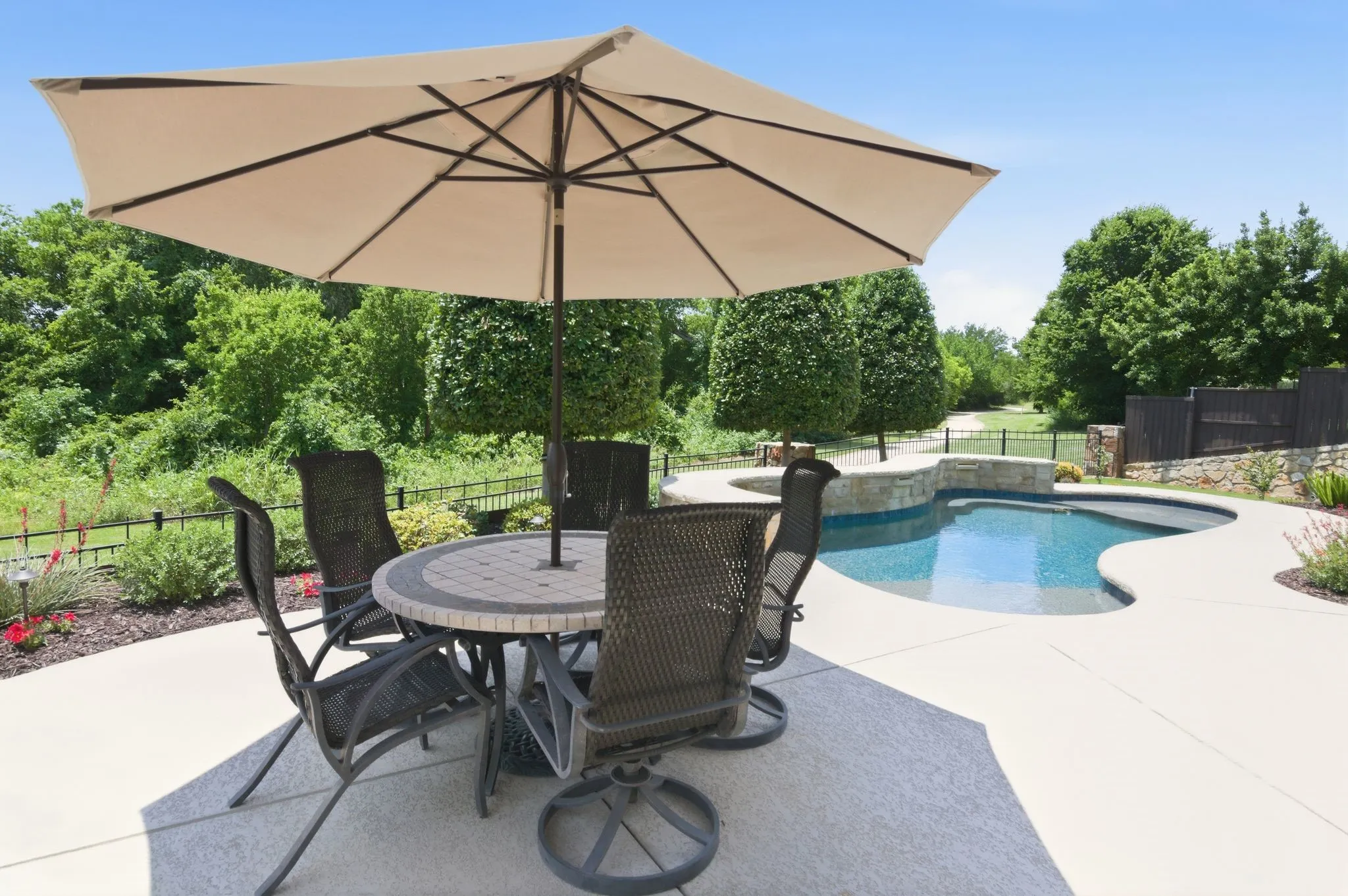 Expansive concrete patio with an integrated swimming pool, surrounded by mature landscaping and a low stone wall