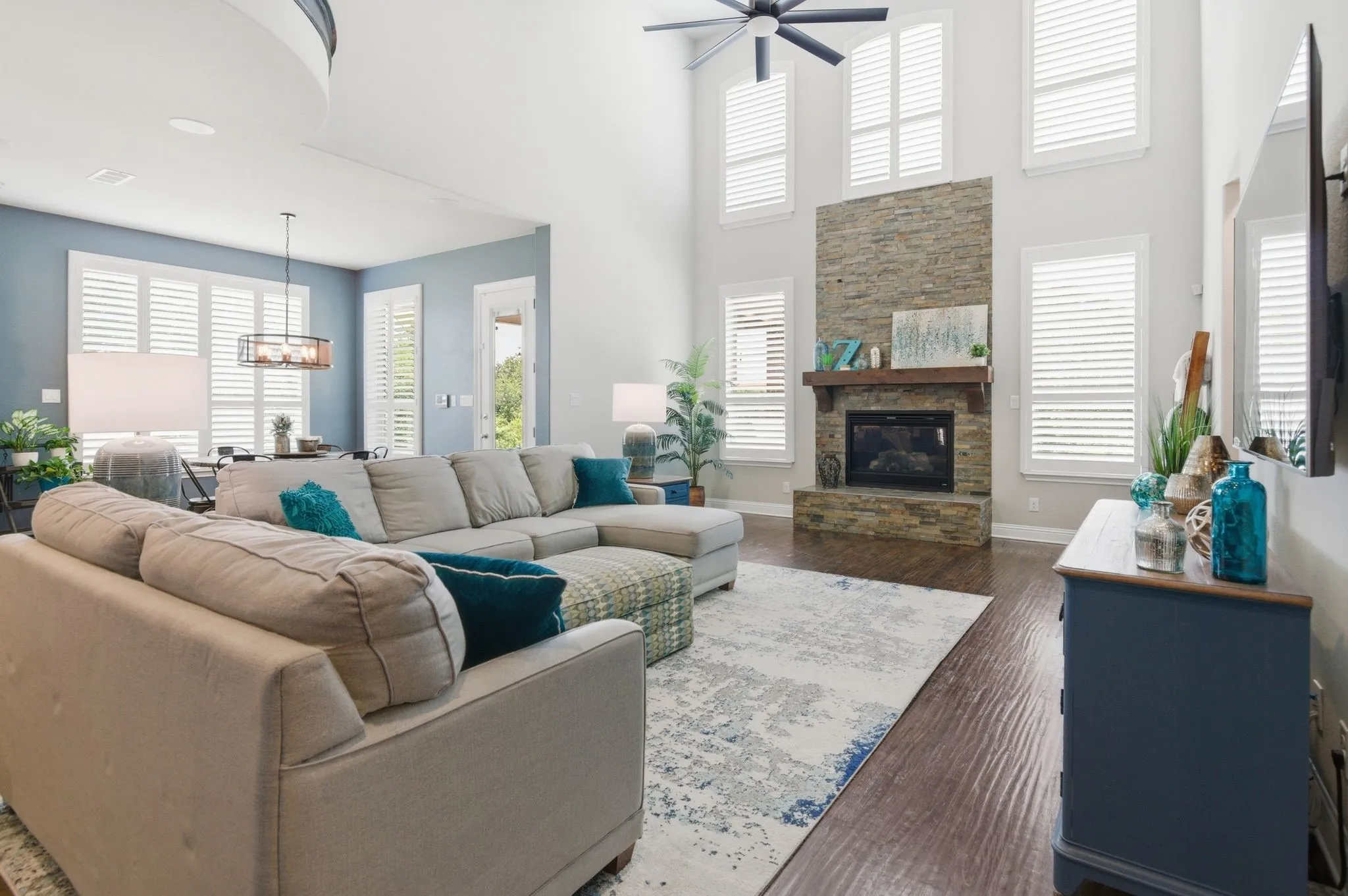 Spacious living area featuring high ceilings, a stacked stone fireplace with a wood mantle, and multiple windows with plantation shutters