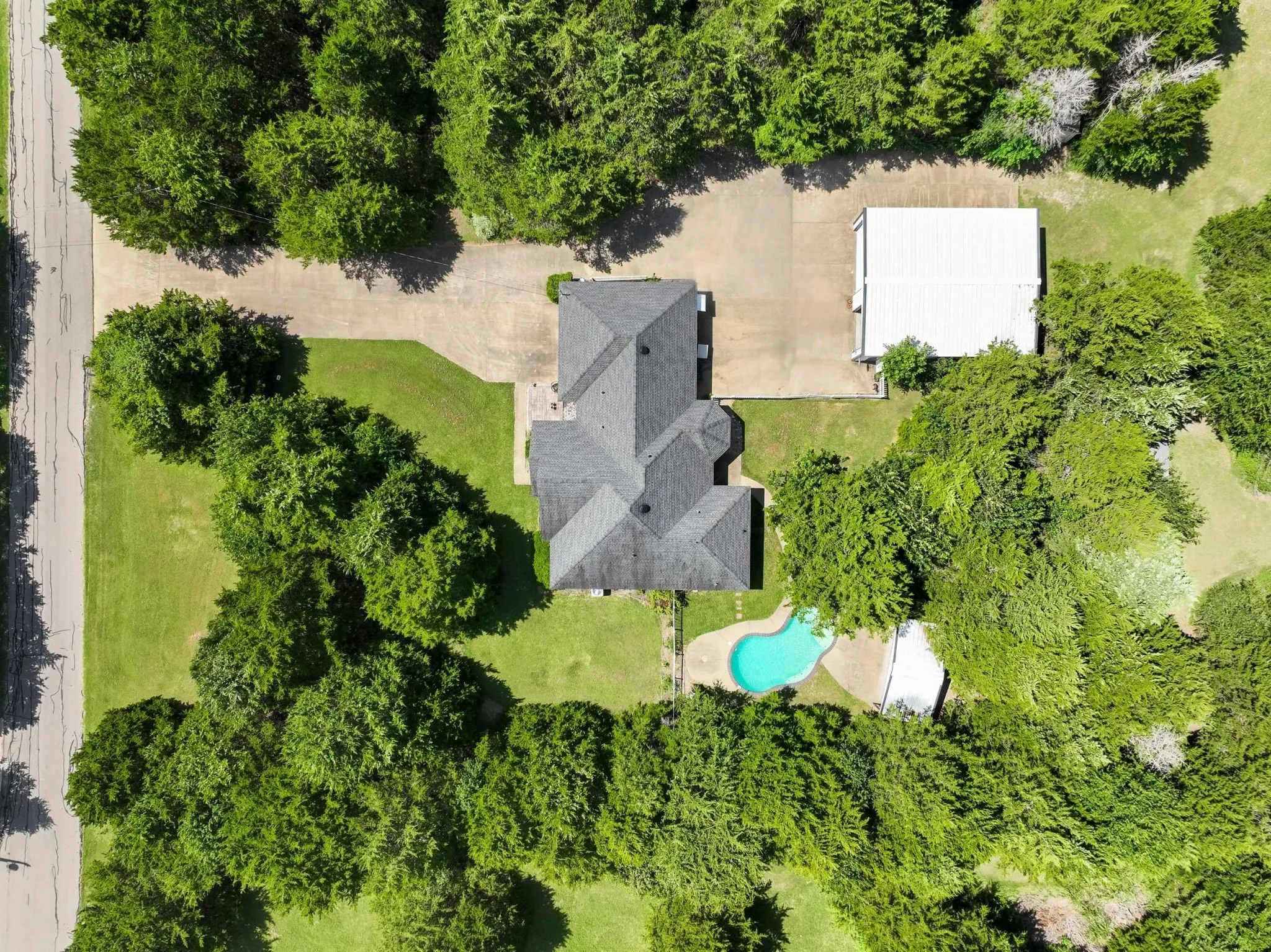 Expansive property featuring a large residence with a dark shingle roof, a detached outbuilding with a light-colored roof, a swimming pool with a stone patio surround, a concrete driveway, and mature landscaping