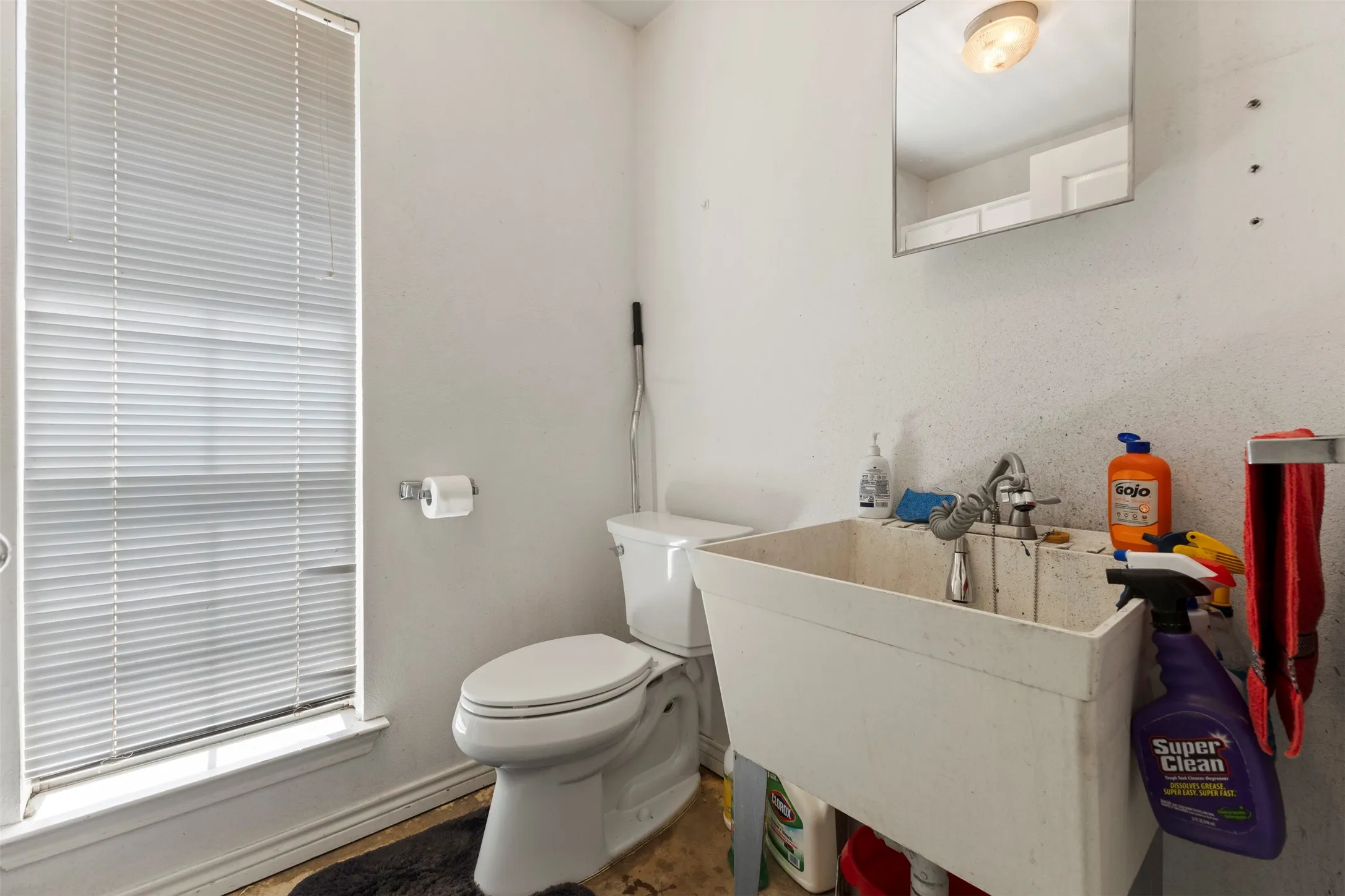 Utility room featuring a window with horizontal blinds, a toilet, a wall-mounted paper holder, a utility sink with faucet, and a wall-mounted mirror