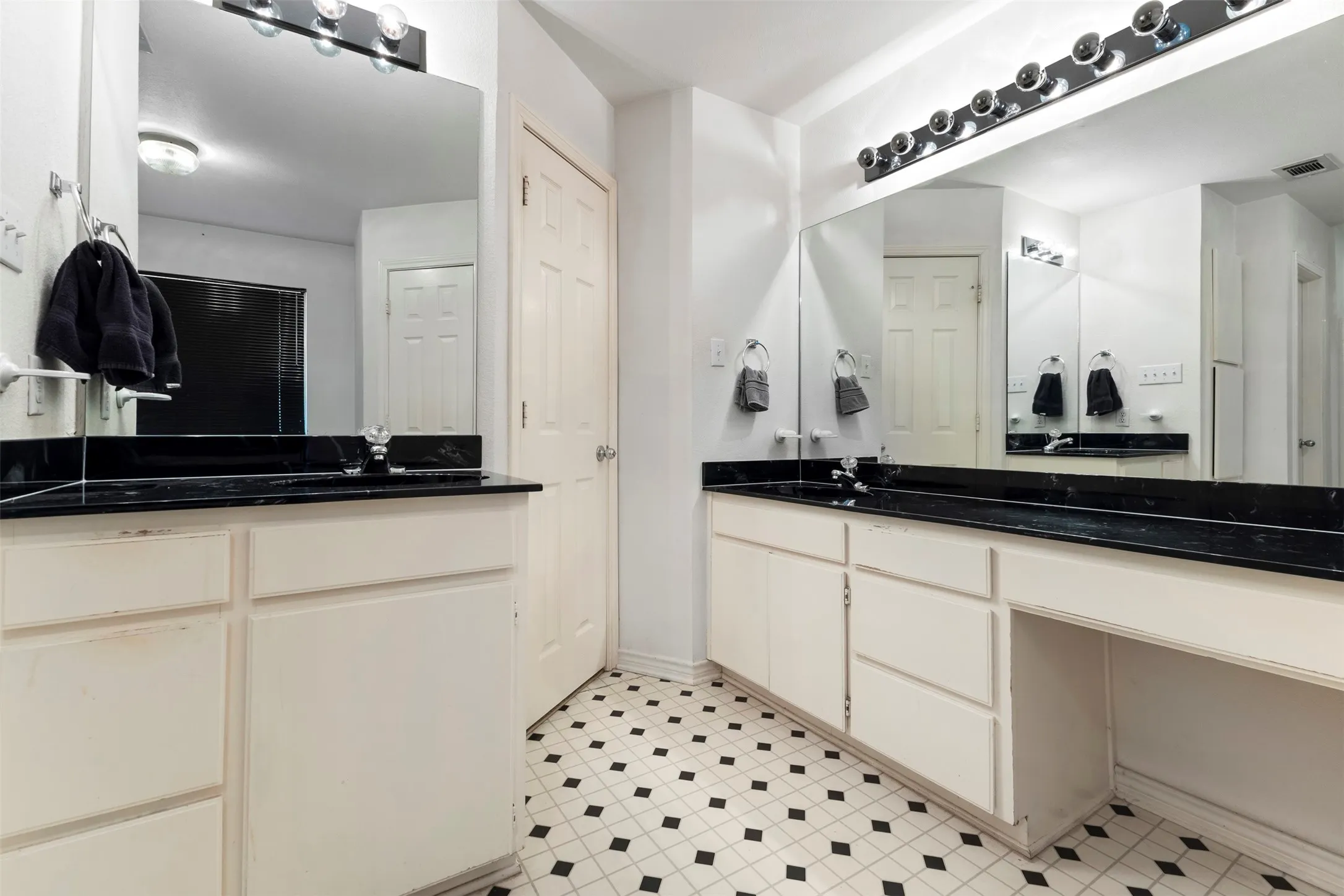 Bathroom featuring dual vanity areas with black countertops, white cabinetry, and a black and white tiled floor