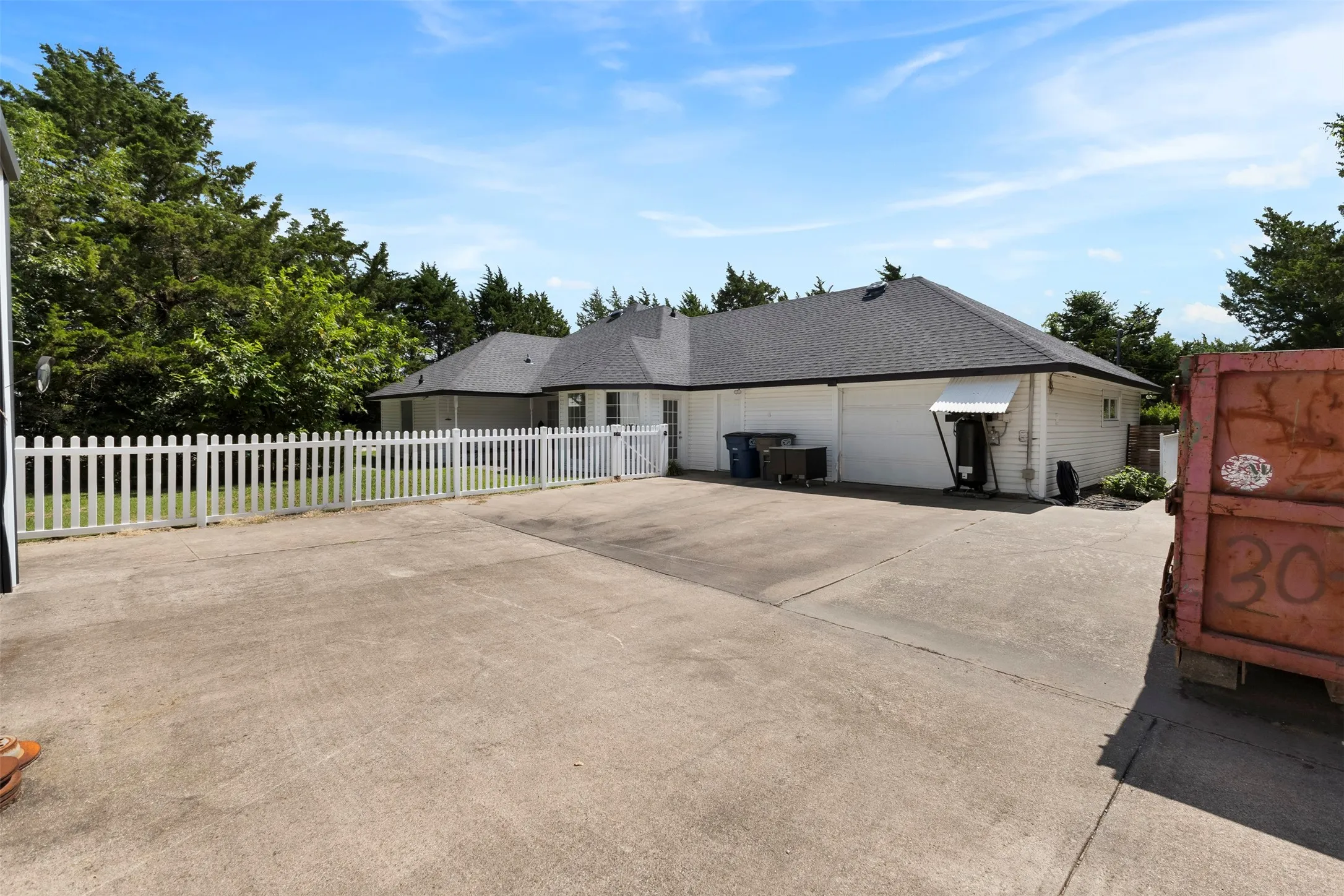 Spacious concrete driveway with a white picket fence, leading to a single-story home featuring a dark shingle roof and white siding