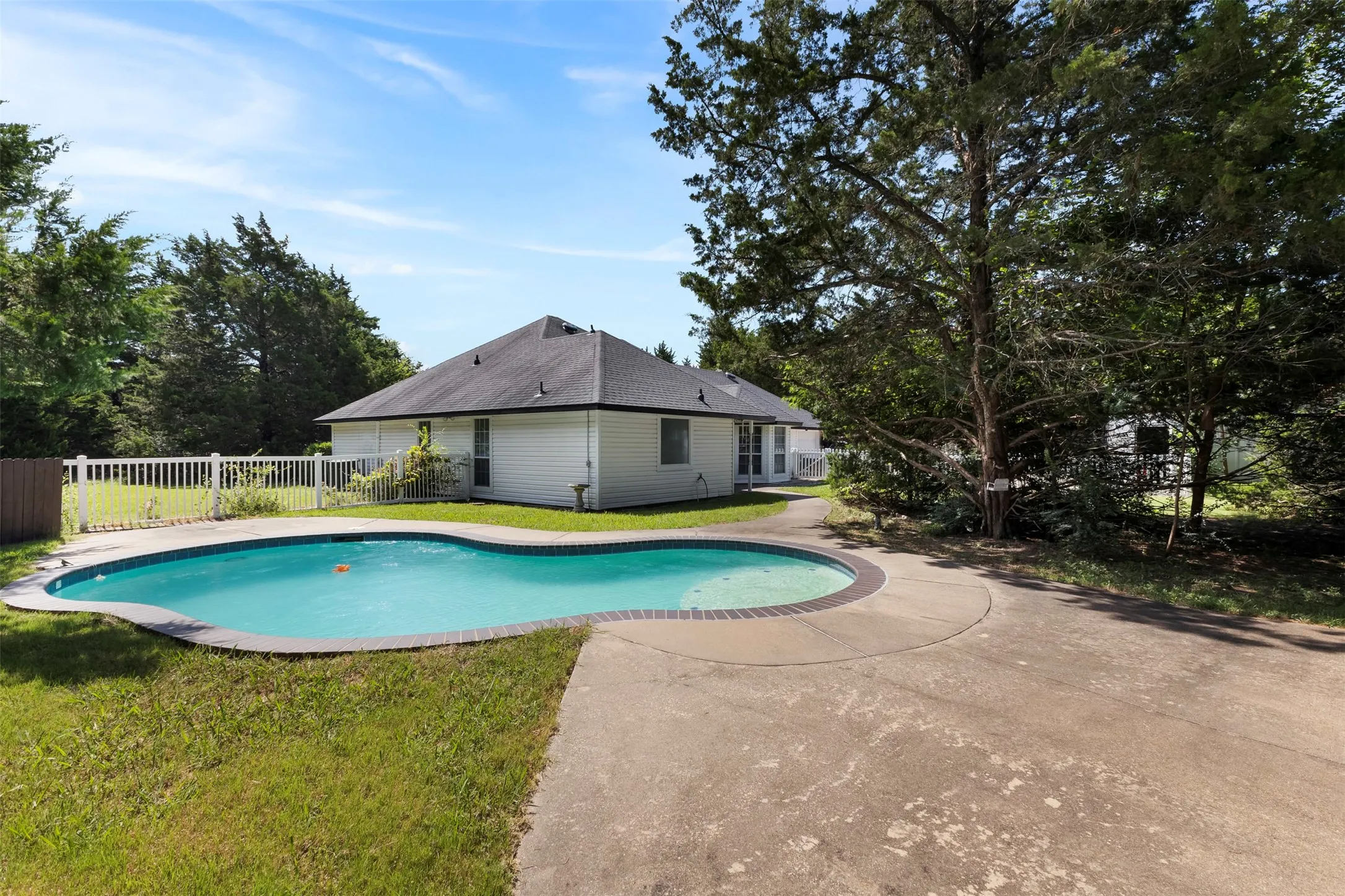 Backyard featuring a free-form swimming pool with concrete decking, surrounded by a white picket fence and mature trees