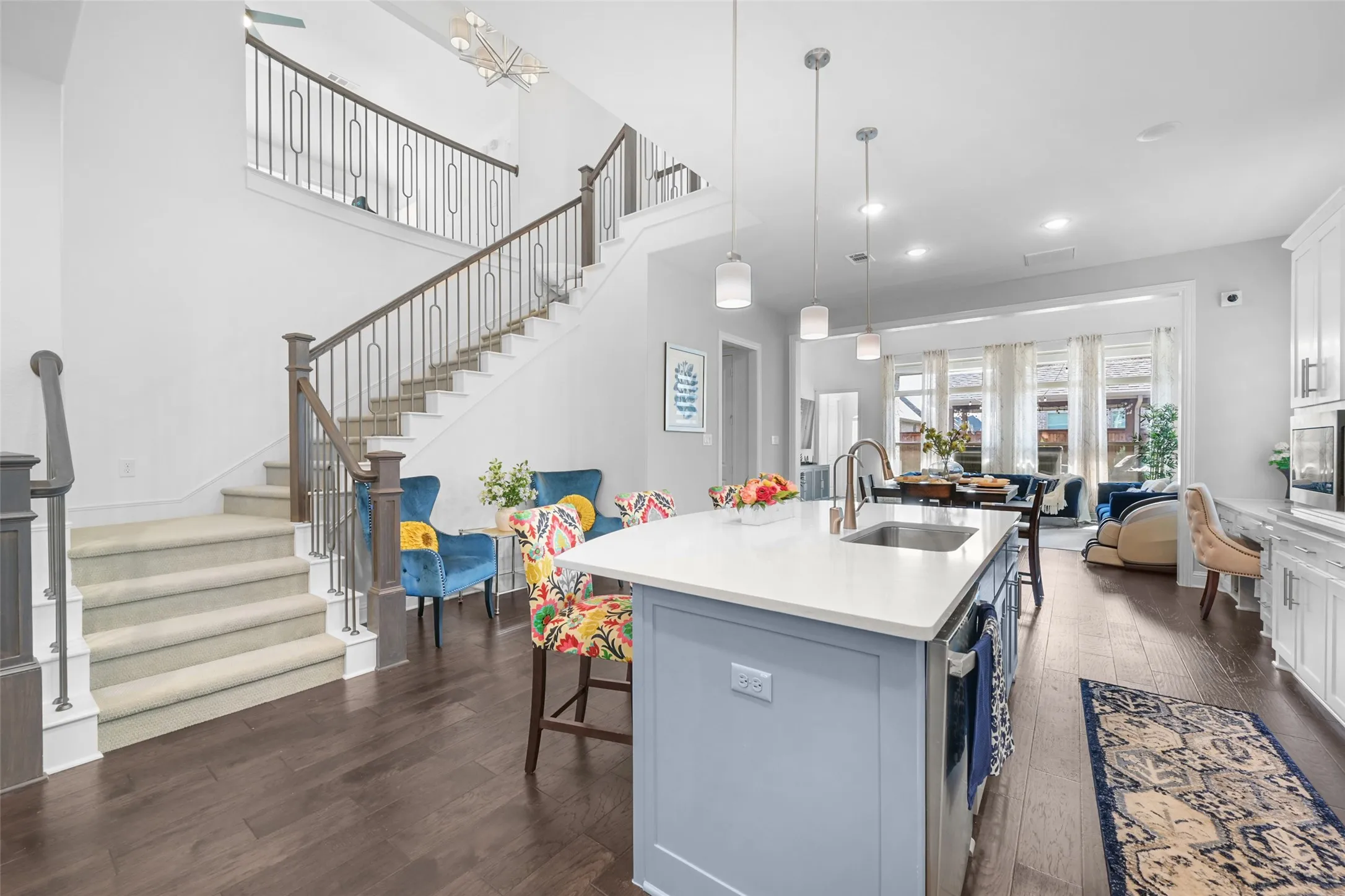 Open-concept living space featuring a two-story entryway, hardwood flooring, a central kitchen island with a sink, white cabinetry, and a carpeted staircase with custom iron balusters