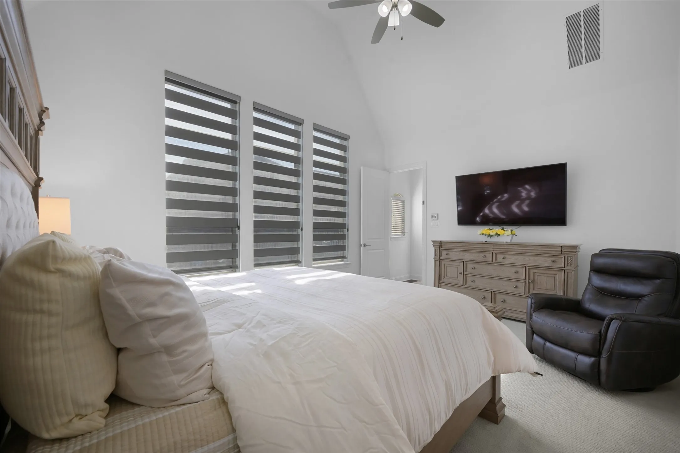 Vaulted ceiling room featuring three elongated windows with horizontal blinds, light-colored carpeting, a ceiling fan, and a built-in wall vent