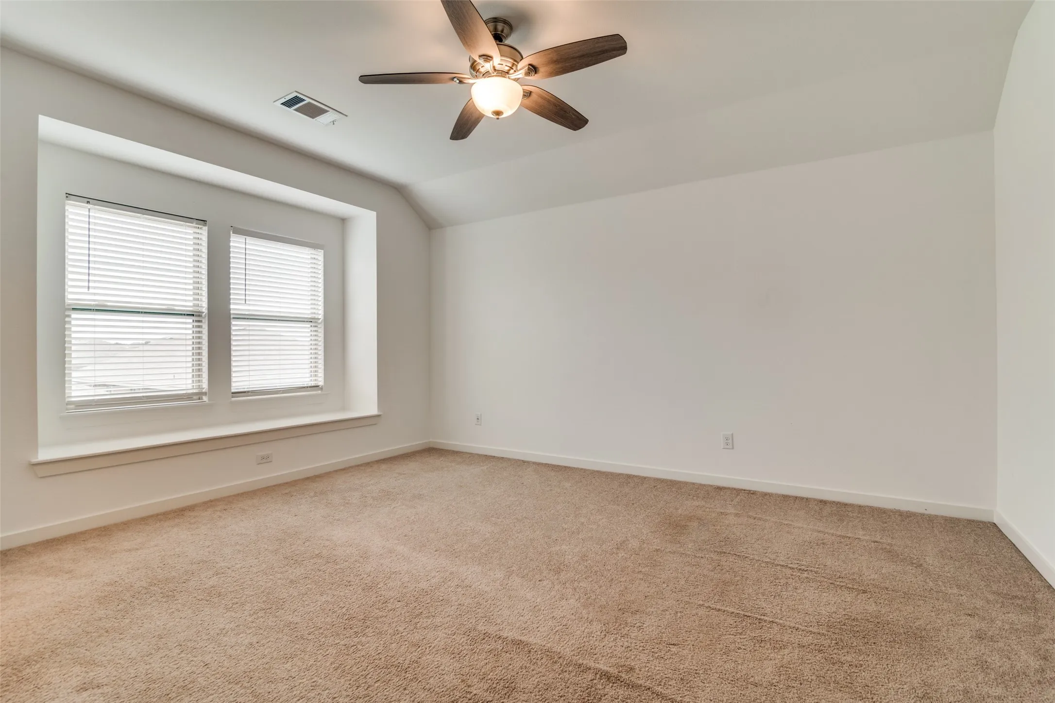 Expansive room featuring a recessed window nook, textured carpeting, a ceiling fan with integrated lighting, pristine white walls, and a sloped ceiling detail