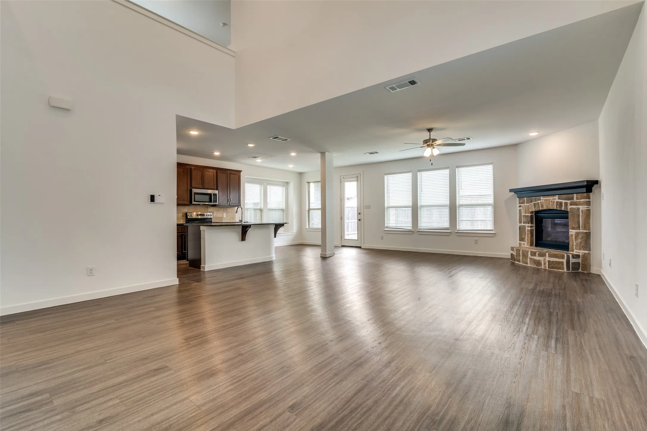 Open-concept interior featuring wood-finish flooring, a stone-faced fireplace with a dark mantel, and a kitchen with dark cabinetry and an island breakfast bar