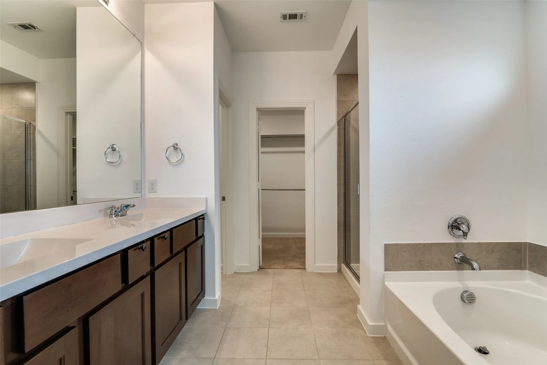Bathroom featuring a dual-sink vanity with a light-colored countertop and wood-finish cabinetry, a white soaking tub, and a glass-enclosed shower