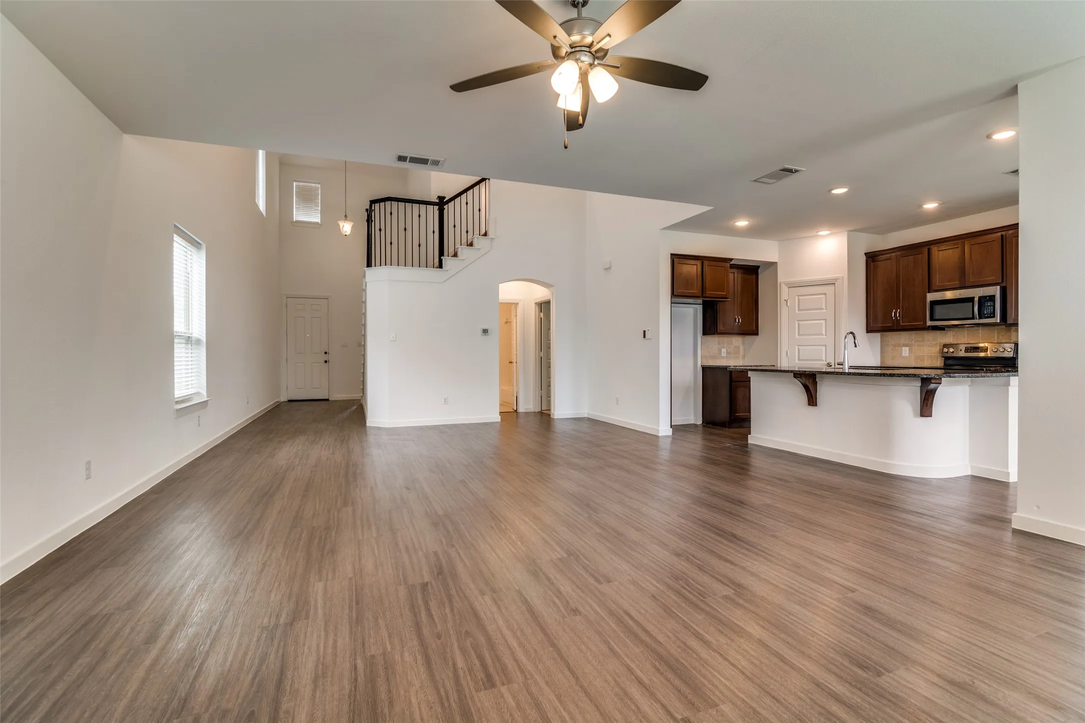 Expansive open-concept room featuring wood-finish flooring, a contemporary ceiling fan, and a kitchen with dark cabinetry, stainless steel appliances, and a breakfast bar with stone countertops