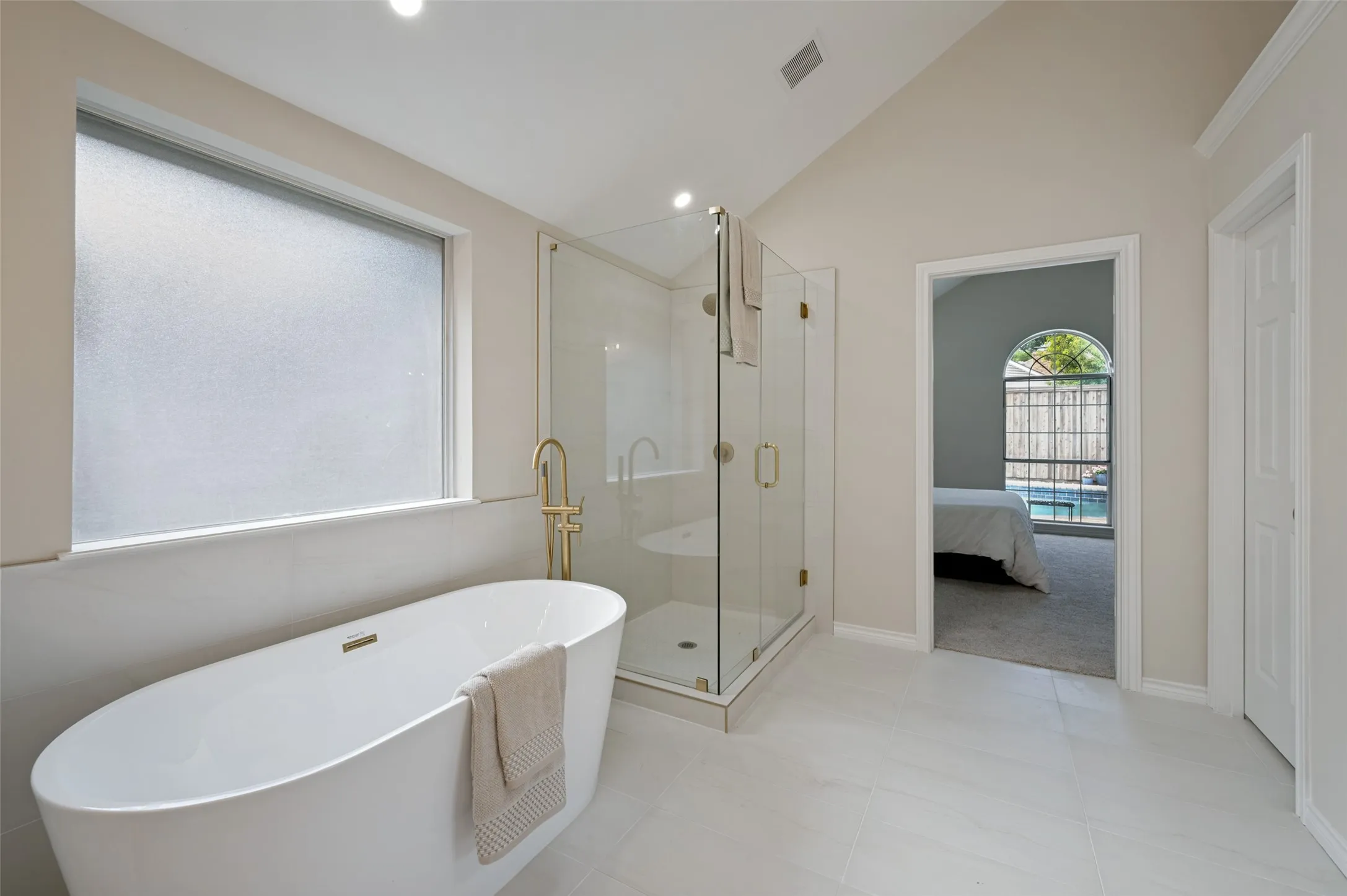 Spacious bathroom featuring a freestanding soaking tub with brass fixtures, a glass-enclosed shower, vaulted ceilings, frosted window, and large format floor tiling